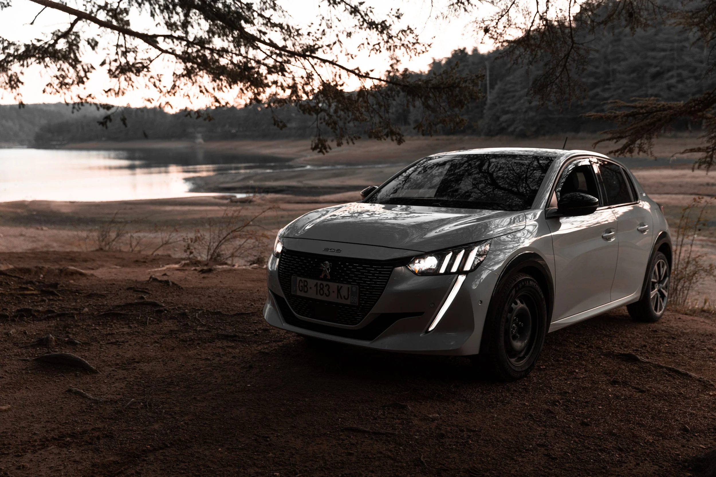 Une voiture blanche stationnée près d'une rivière dans un environnement naturel, avec des arbres en arrière-plan et un ciel de crépuscule.