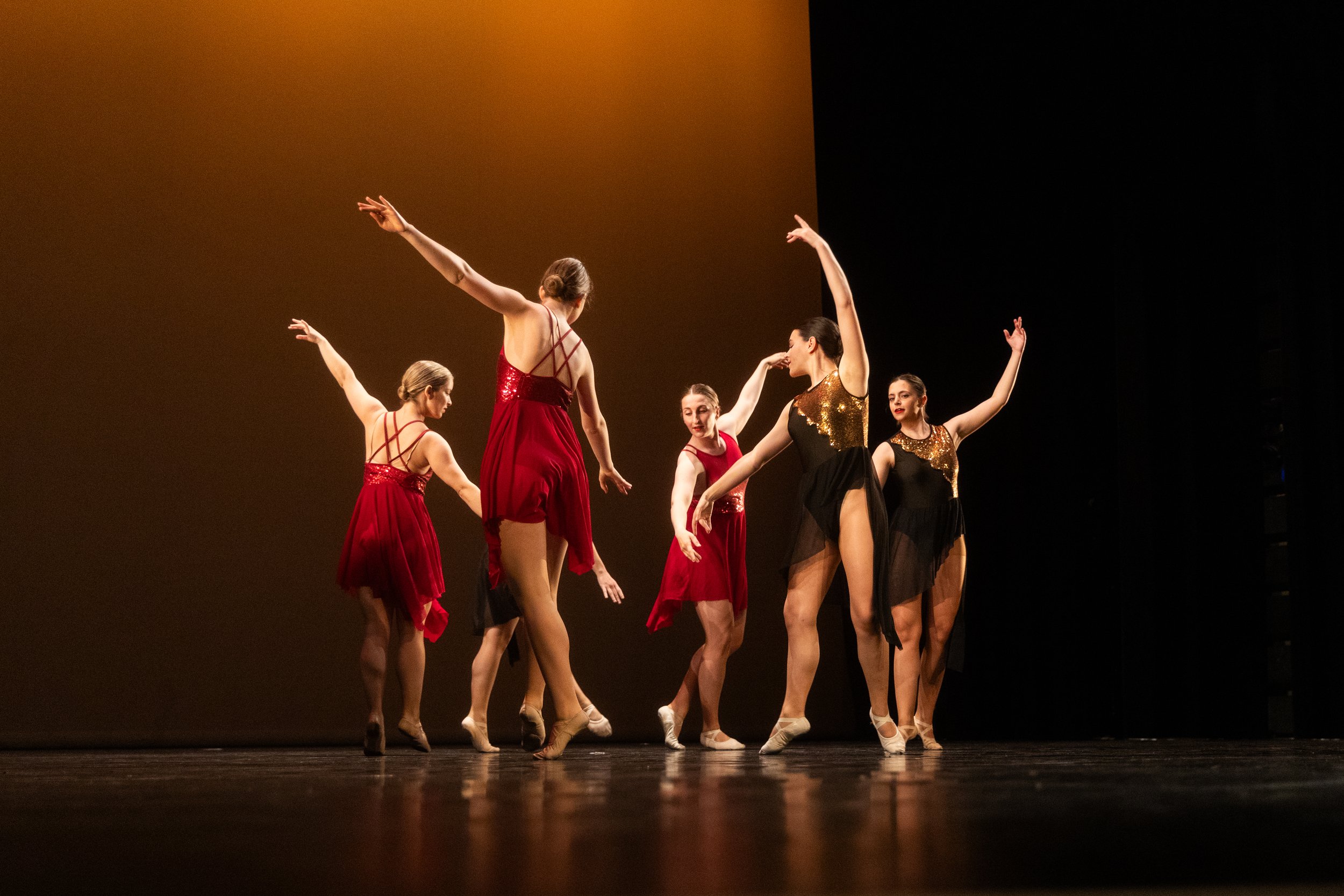 Six ballerinas en costumes colorés réalisant une performance de danse sur scène avec une lumière sobre.