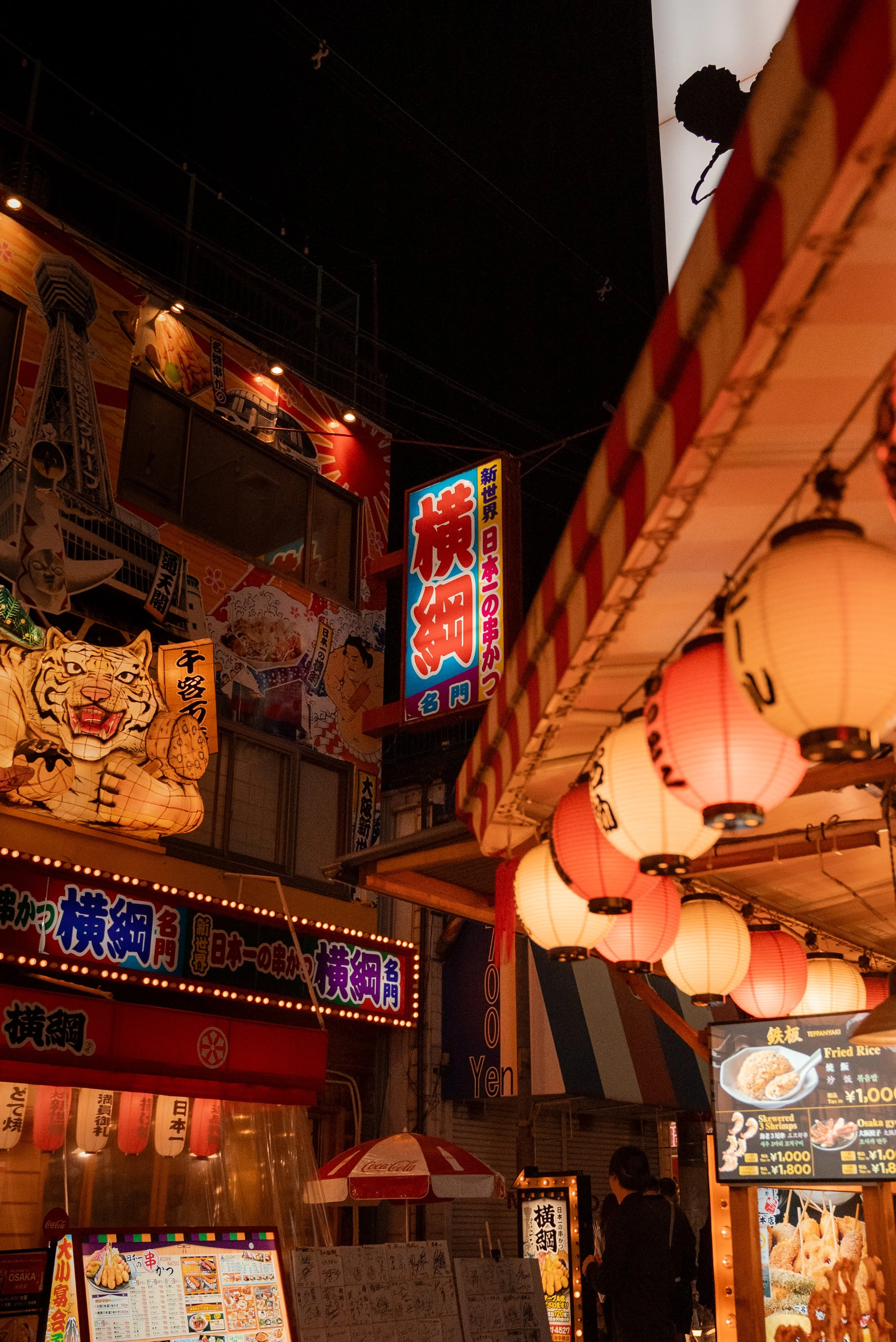 Une scène de nuit dans une rue commerciale au Japon avec des enseignes lumineuses en japonais, des lanternes traditionnelles, et des décorations colorées, y compris un tigre en papier lanternes et des menus de nourriture.