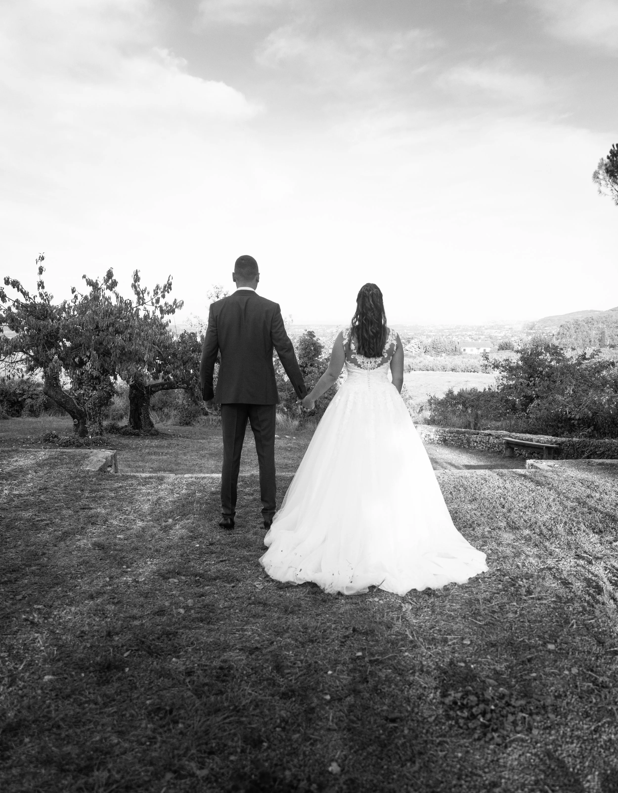 Un couple en tenue de mariage se tient la main, vue de dos, dans un paysage extérieur avec des arbres et un ciel nuageux.