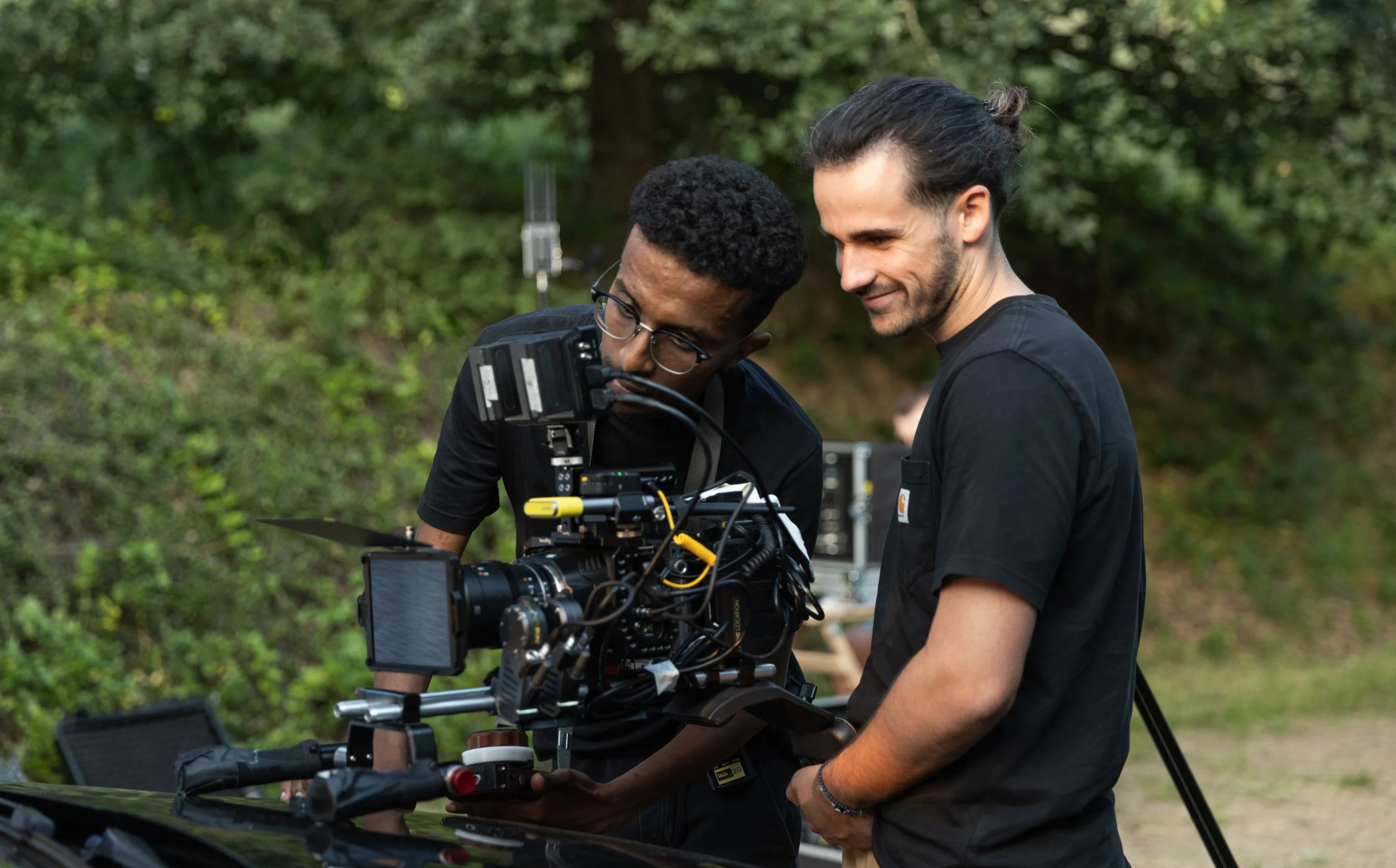 Deux hommes, un avec des lunettes et un autre avec les cheveux attachés, travaillent sur une caméra de tournage à l'extérieur, au milieu de la nature.