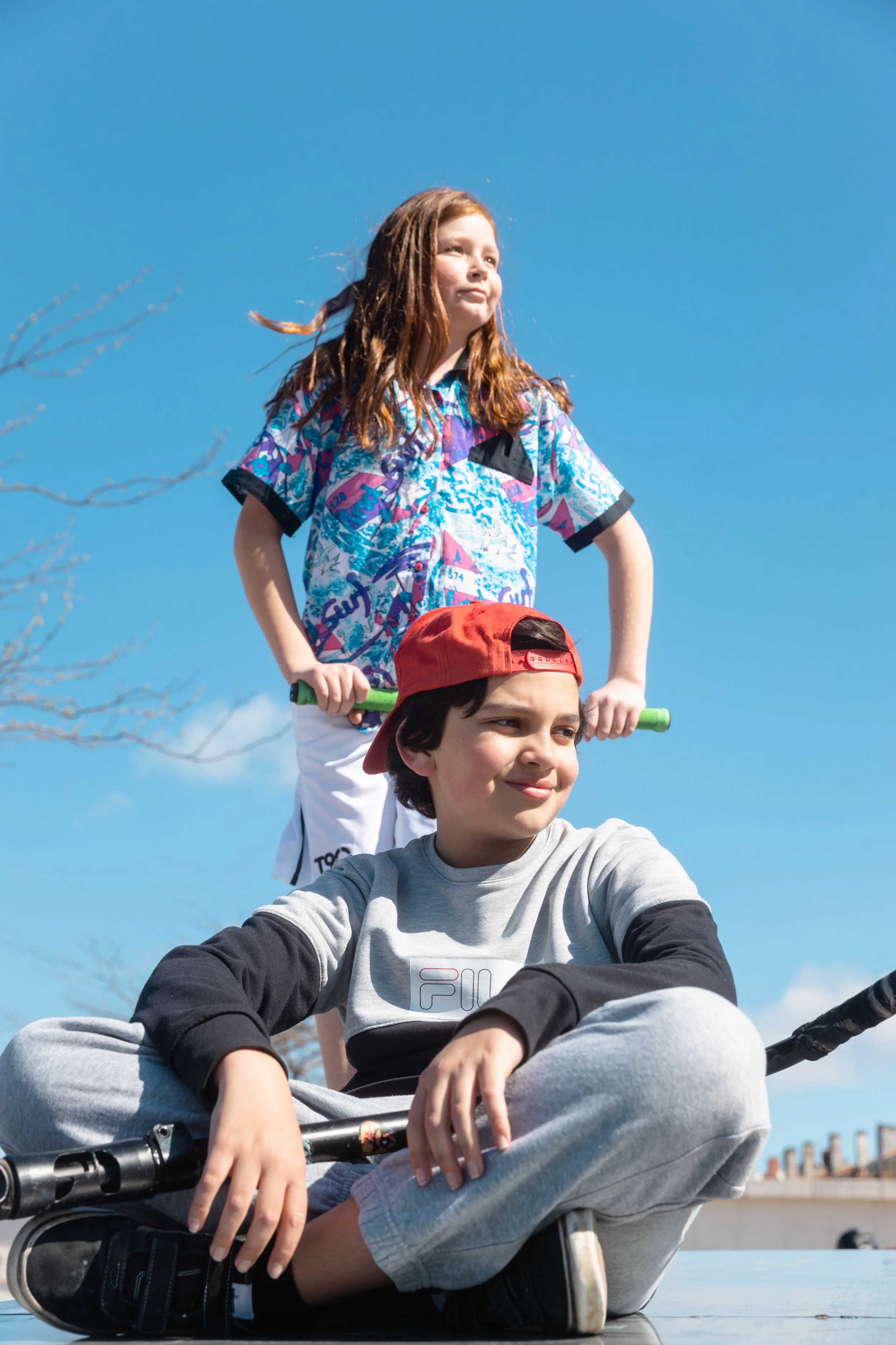 Un garçon assis sur un skate, portant un sweat à capuche gris avec des manches noires, une casquette rouge à l'envers, et tenant un scooter, avec une fille derrière lui qui pousse le scooter, tous deux dehors sous un ciel bleu.