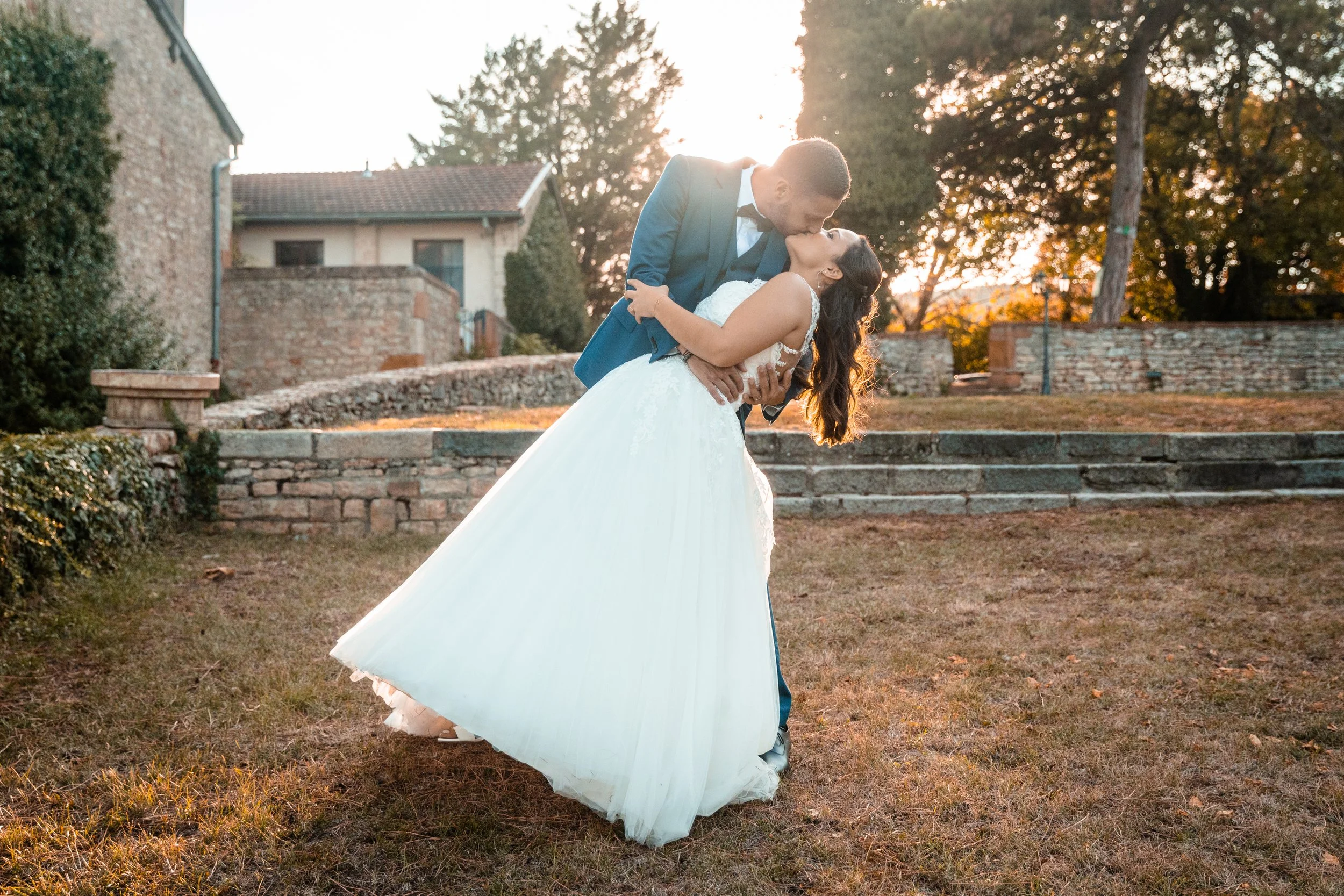 Un couple en costume de mariage, l'homme portant un costume bleu et la femme en robe de mariée blanche, partage un baiser dans un jardin au coucher du soleil.