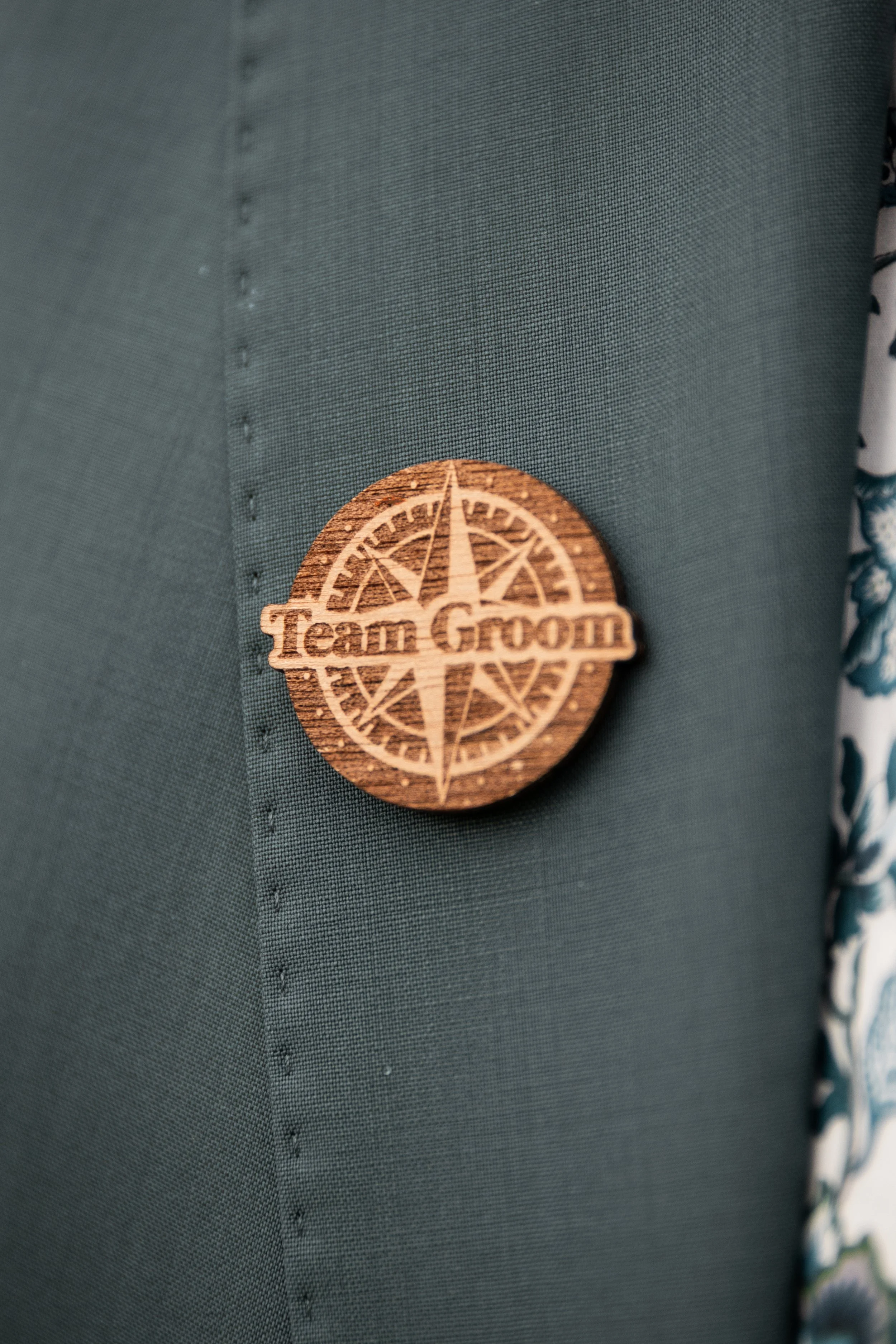 Un badge en bois gravé avec une boussole et le texte 'Team GROOM', placé sur un tissu vert. À droite, une partie d'un tissu à motif floral.