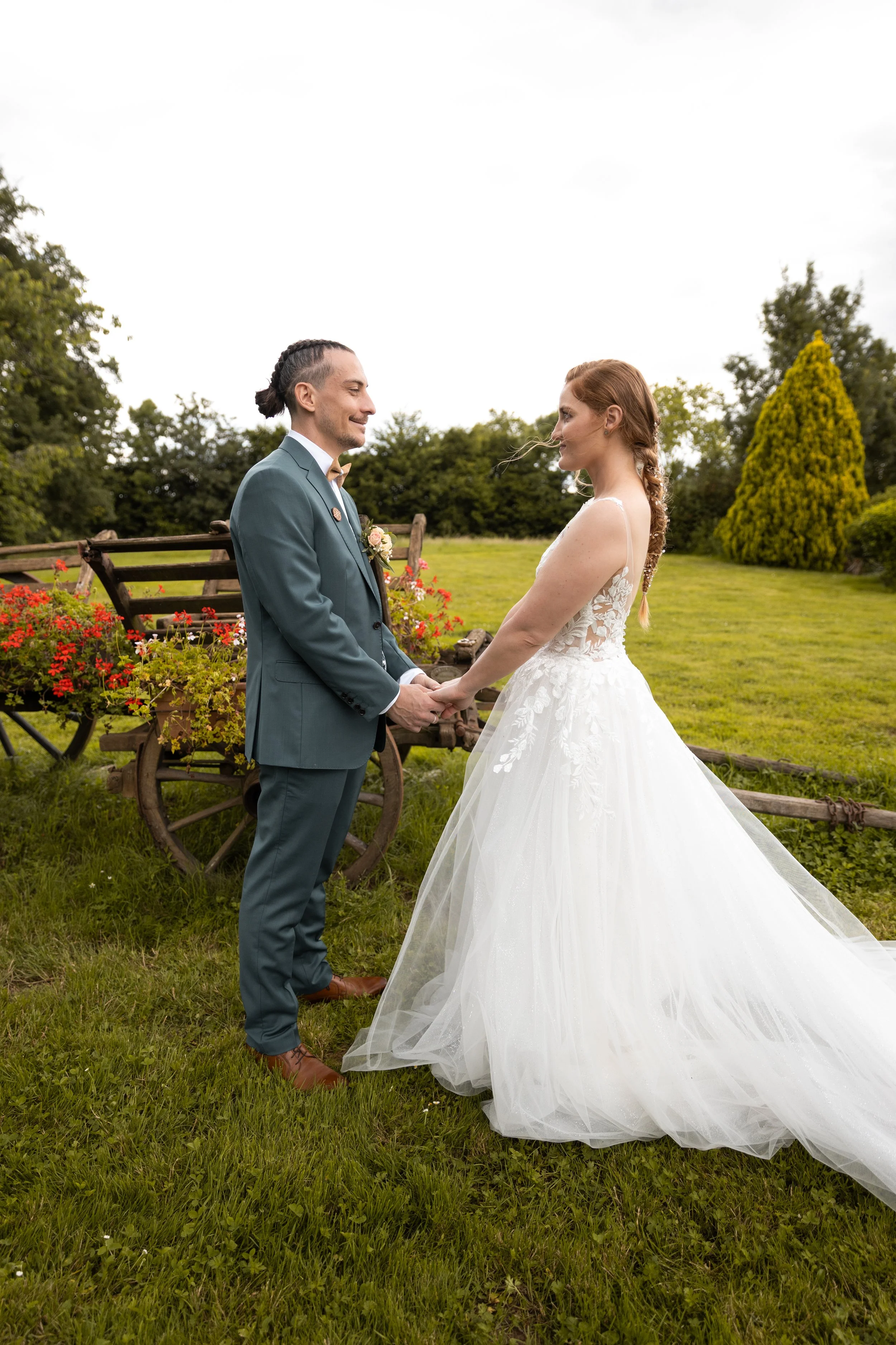 Un couple lors d'un mariage, la mariée en robe blanche et le marié en costume bleu, se tenant la main dans un jardin vert avec des fleurs et des arbres.