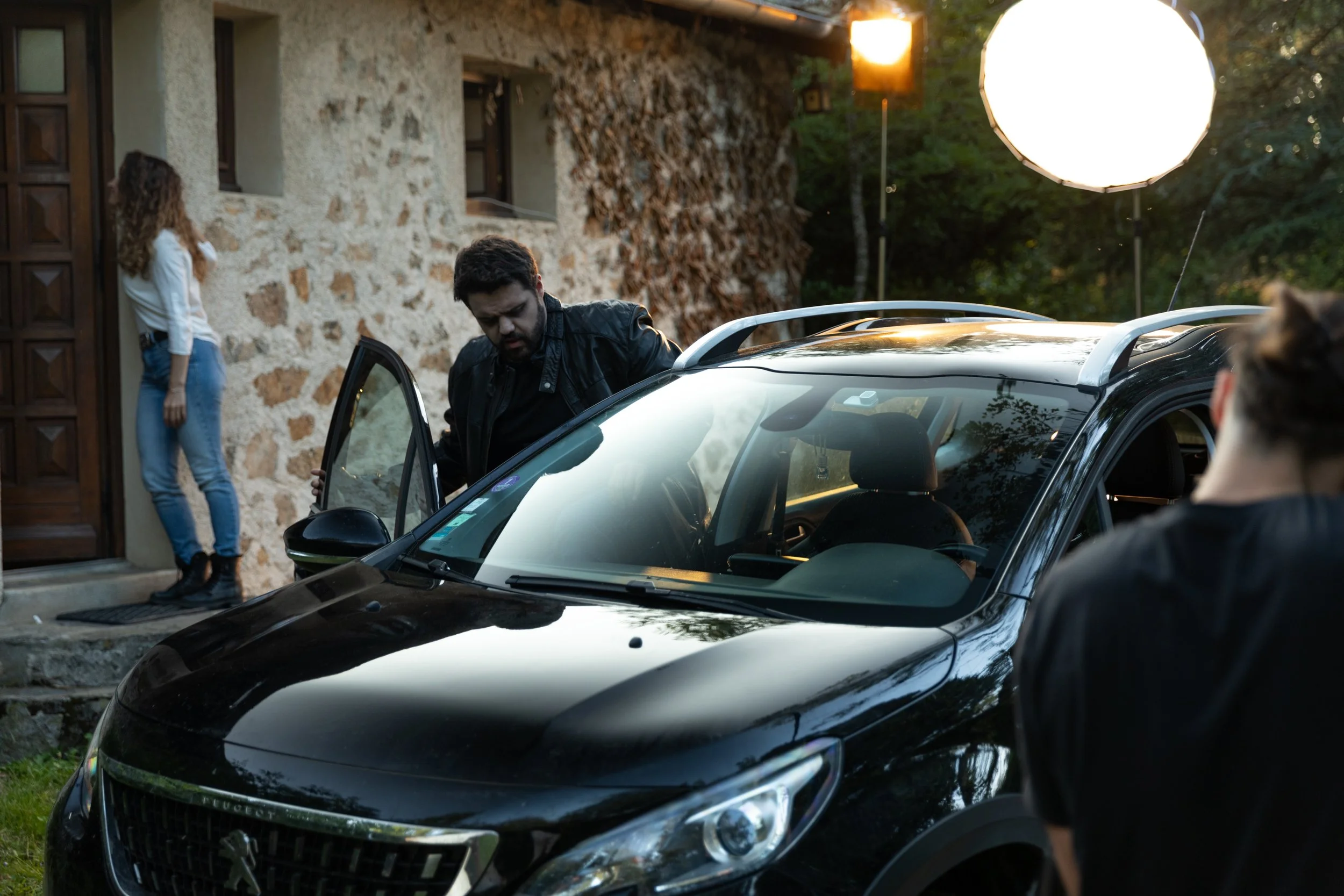 Une équipe de tournage devant une vieille maison en pierre, avec un véhicule noir et un éclairage professionnel, filmant une scène extérieure en fin d'après-midi ou début de soirée.