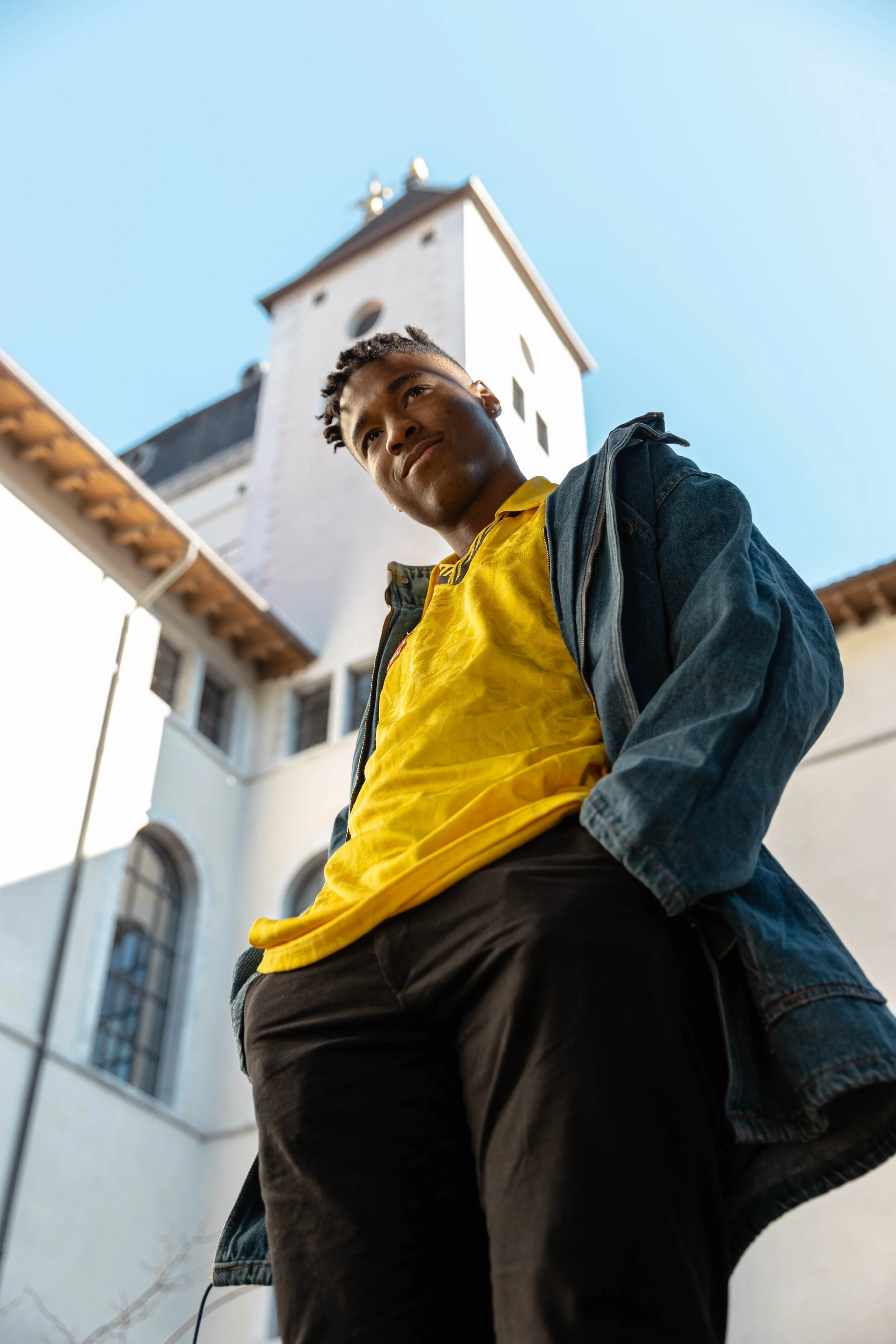 Jeune homme regardant vers le haut, vêtement jaune et veste en jean, bâtiment blanc au fond, ciel clair.