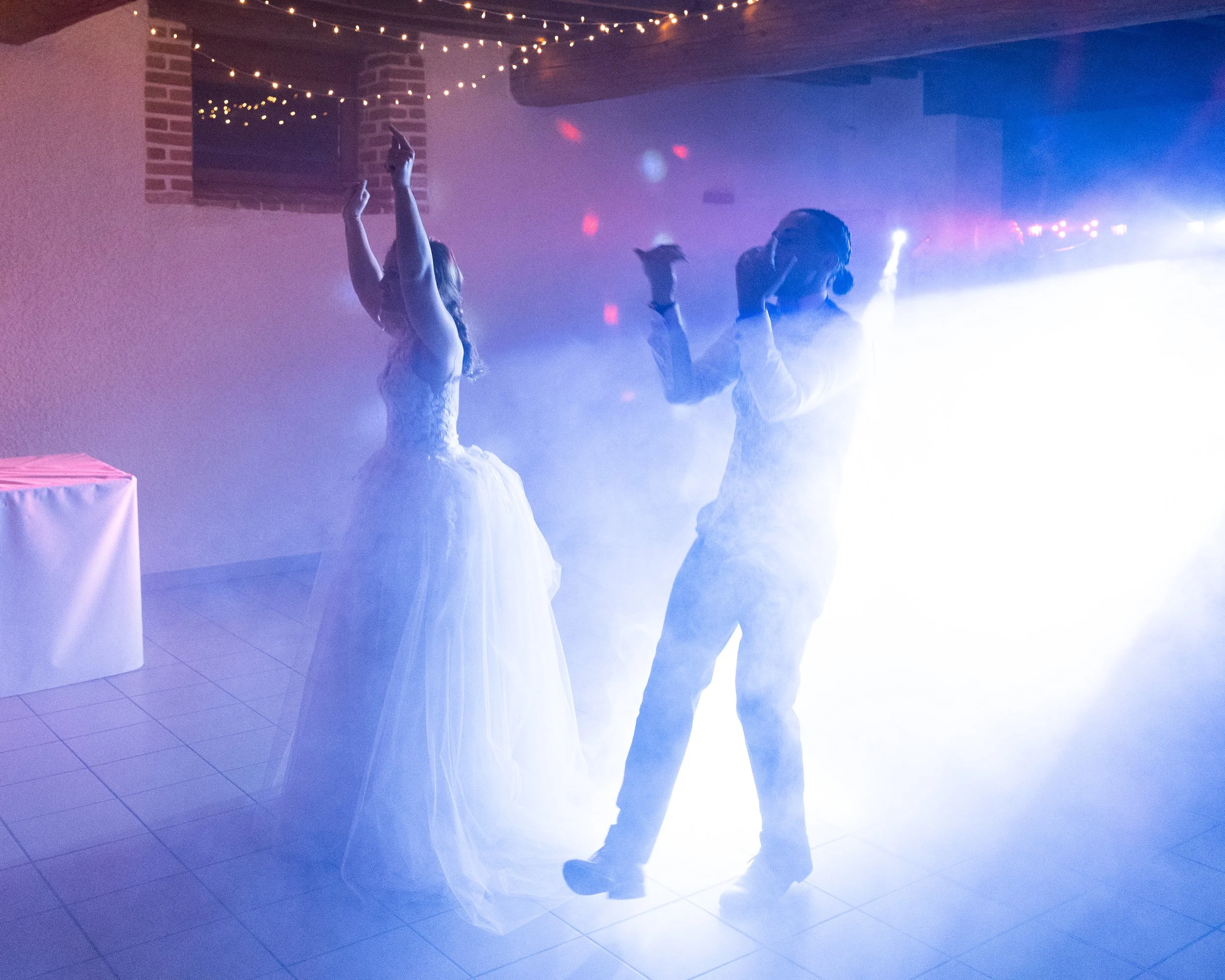 Une mariée et un marié dansent lors d'une réception de mariage, entourés de fumée et éclairés par des lumières colorées dans une salle décorée avec des guirlandes lumineuses.