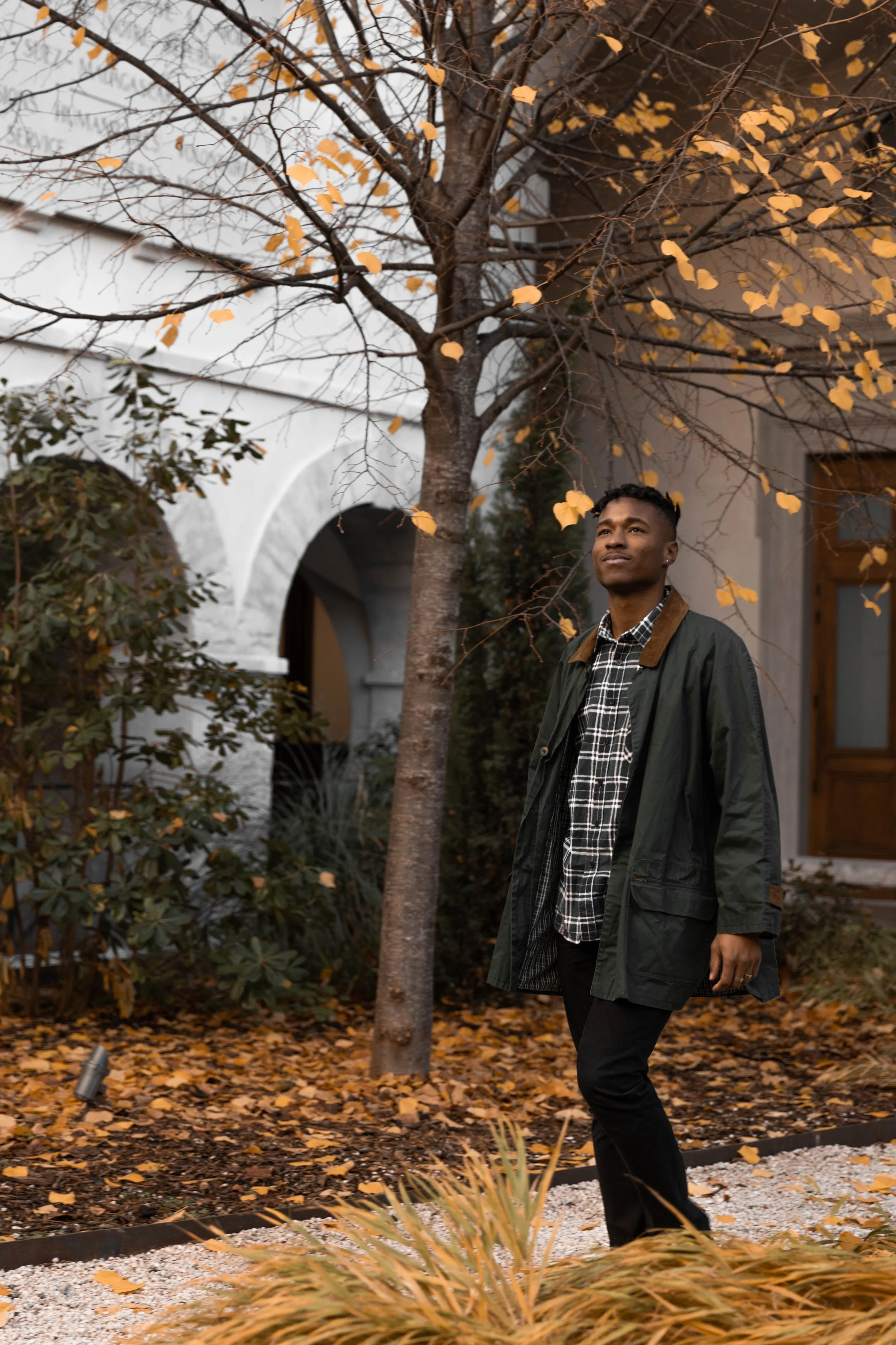 Un homme marche dans un jardin aux feuilles d'automne, portant une veste verte et un chemisier à carreaux noir et blanc, avec un bâtiment blanc en arrière-plan.