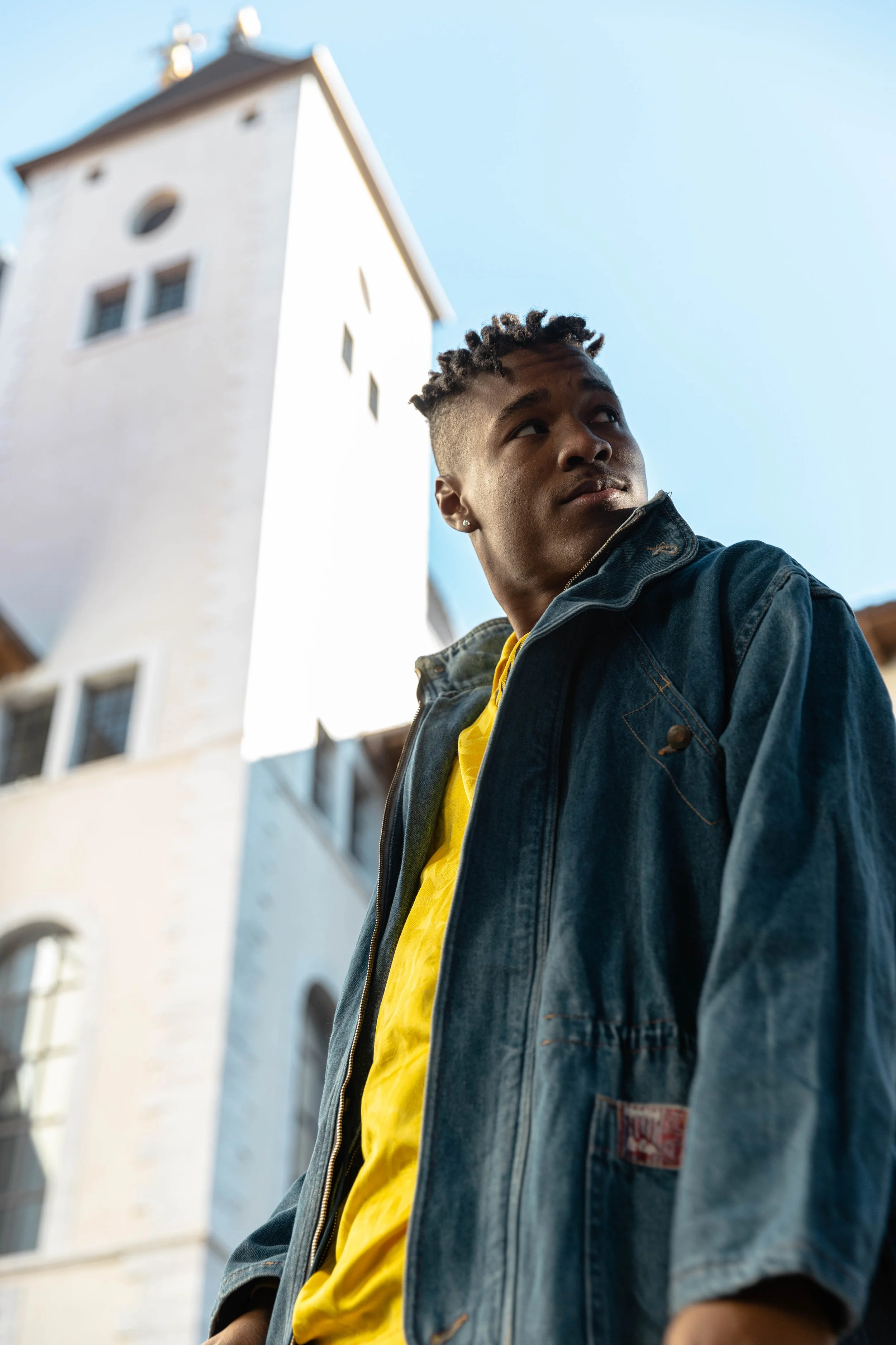 Jeune homme regardant vers le haut, portant une veste en denim et un t-shirt jaune, avec une grande tour blanche en arrière-plan sous un ciel bleu.