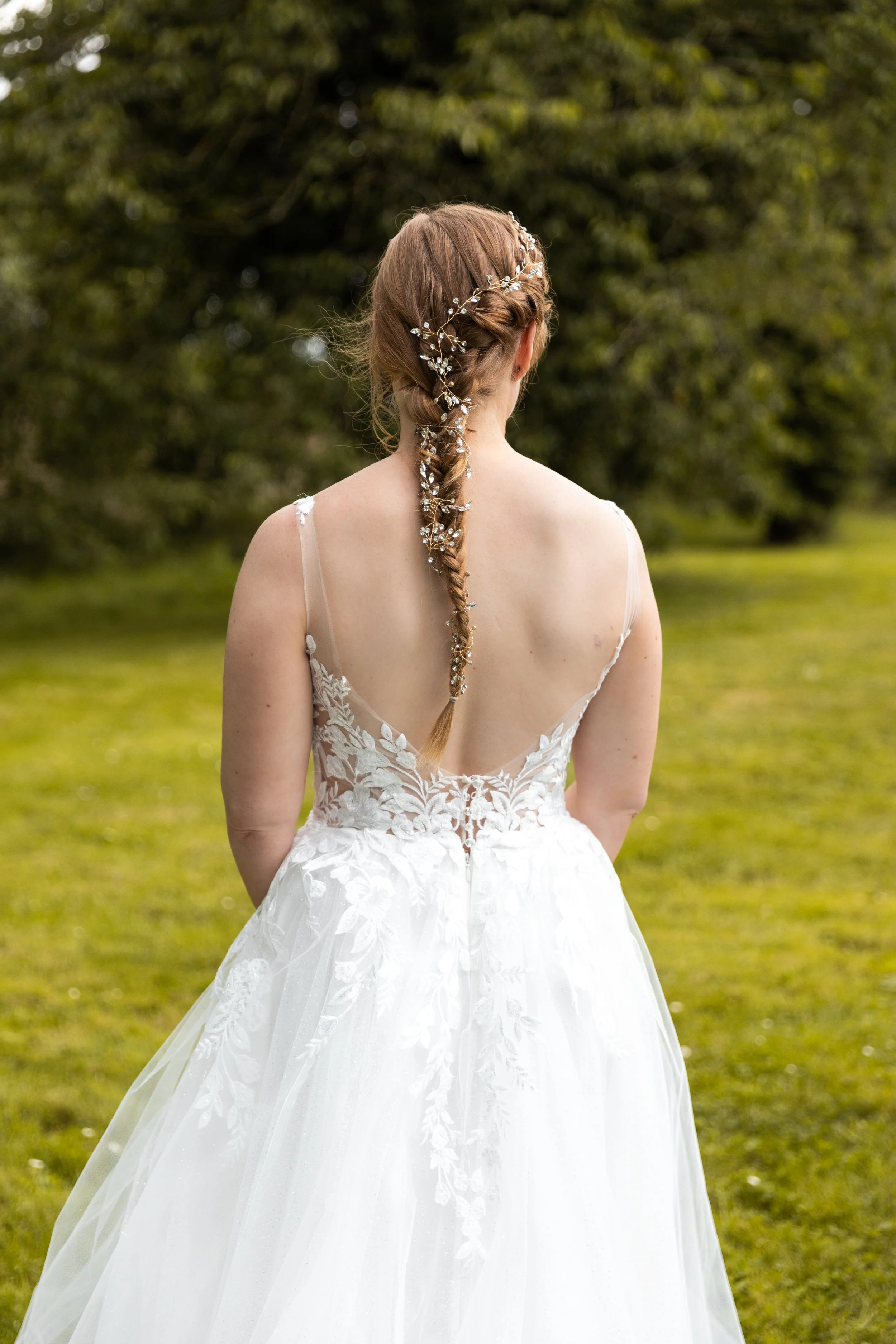 Une femme en robe de mariée avec ses cheveux attachés à l'arrière, portant une couronne de fleurs et de perles, vue de dos, dans un jardin verdoyant.