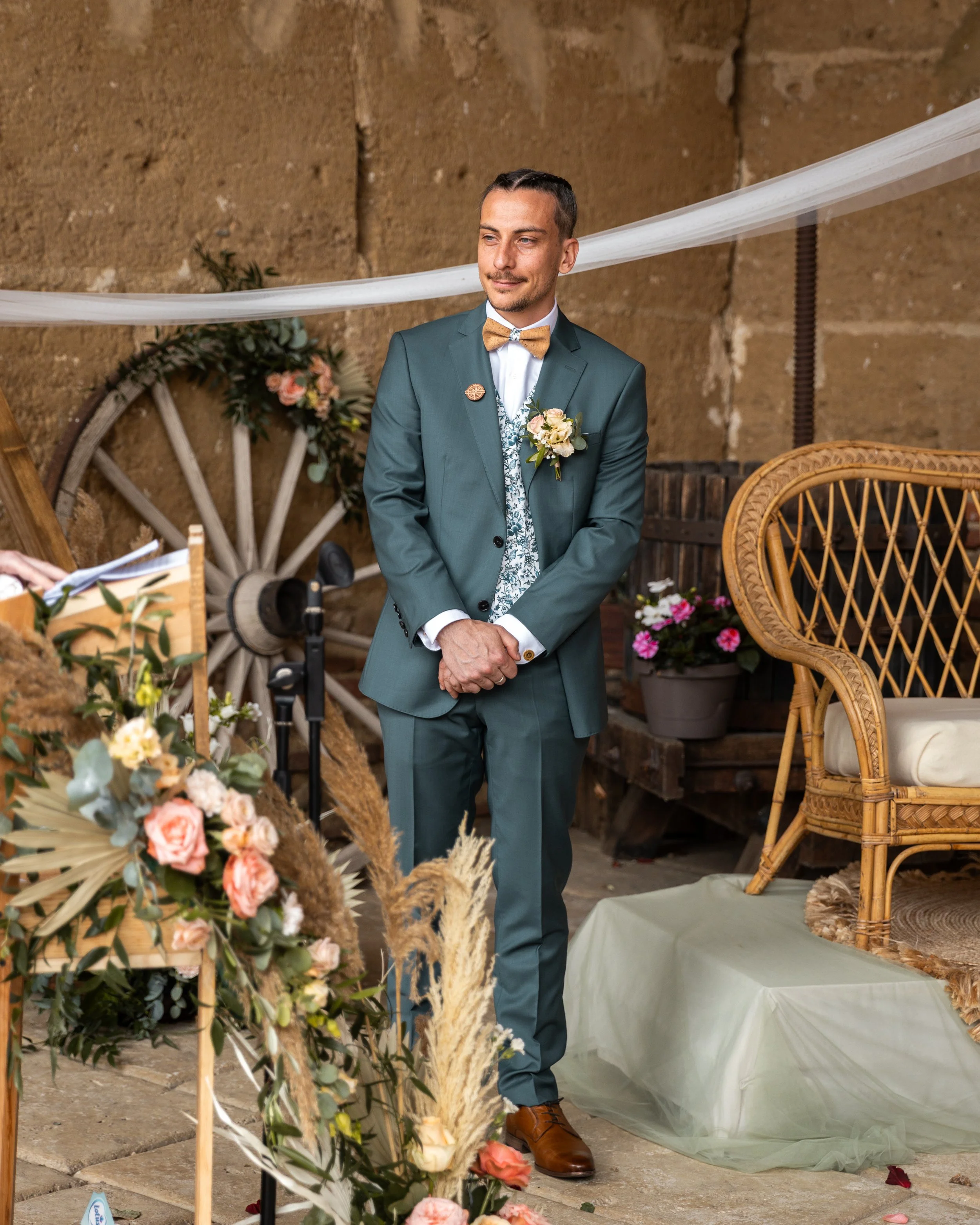 Homme lors d'une cérémonie de mariage, portant un costume vert avec une chemise blanche, un nœud papillon beige, une boutonnière de fleurs et se tenant parmi des décorations florales et champêtres dans un lieu rustique.