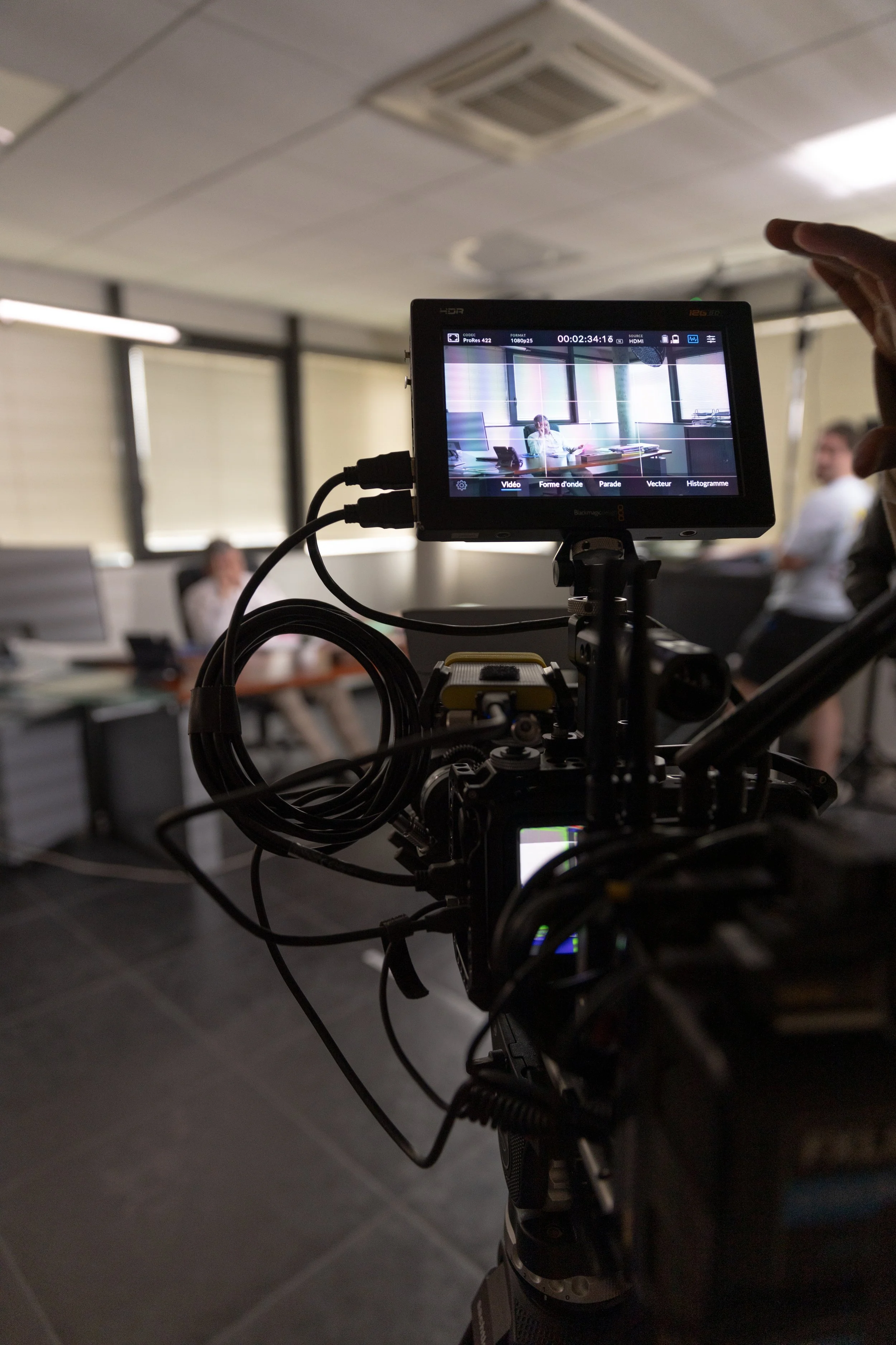 Une caméra filme une réunion dans une salle de conférence avec trois personnes, une assise à une table et deux debout, dans une pièce bien éclairée par des fenêtres.