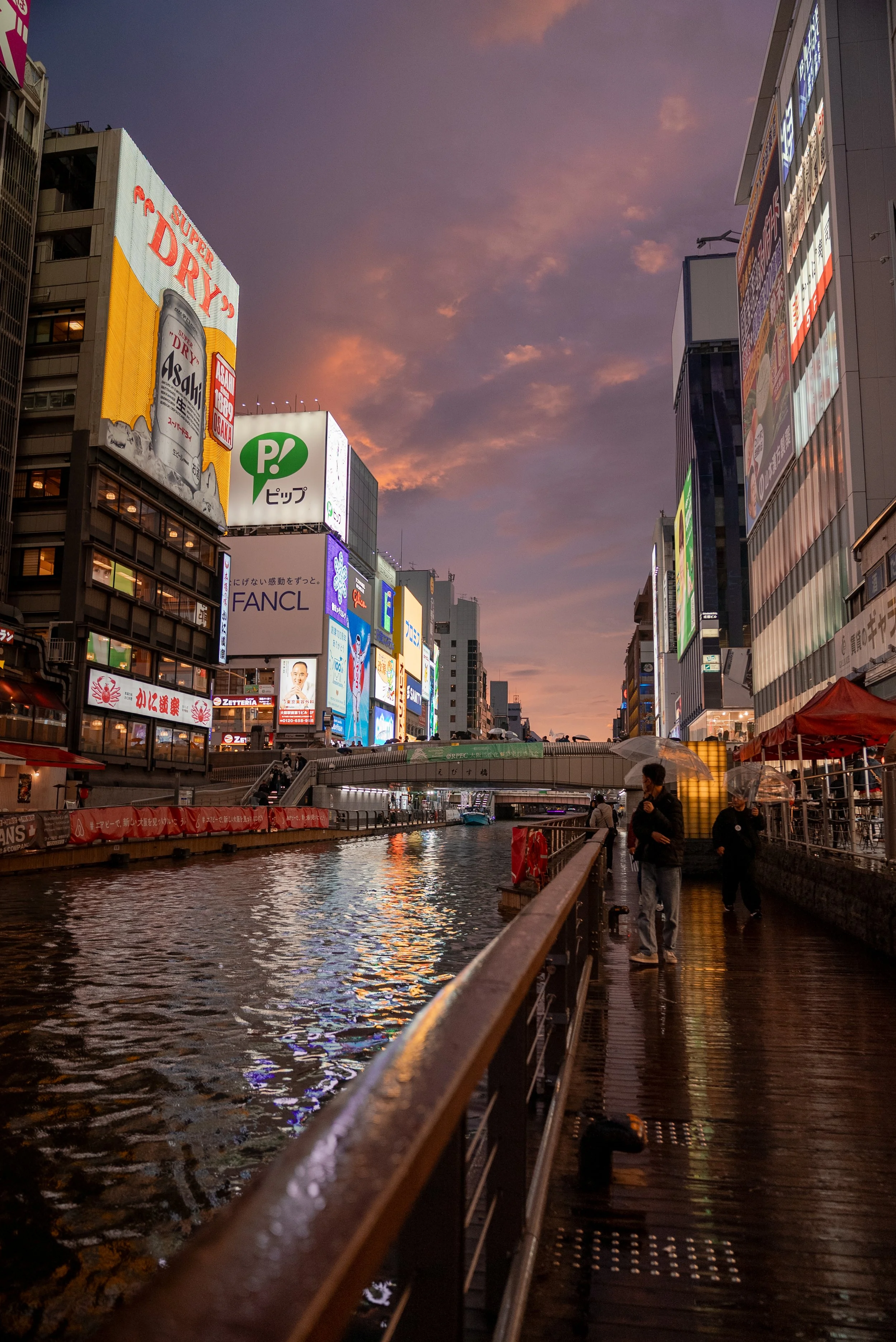 Vue d'une ville japonaise au crépuscule, avec des bâtiments illuminés et un canal réfléchissant les lumières, des personnes marchant sous des parapluies.