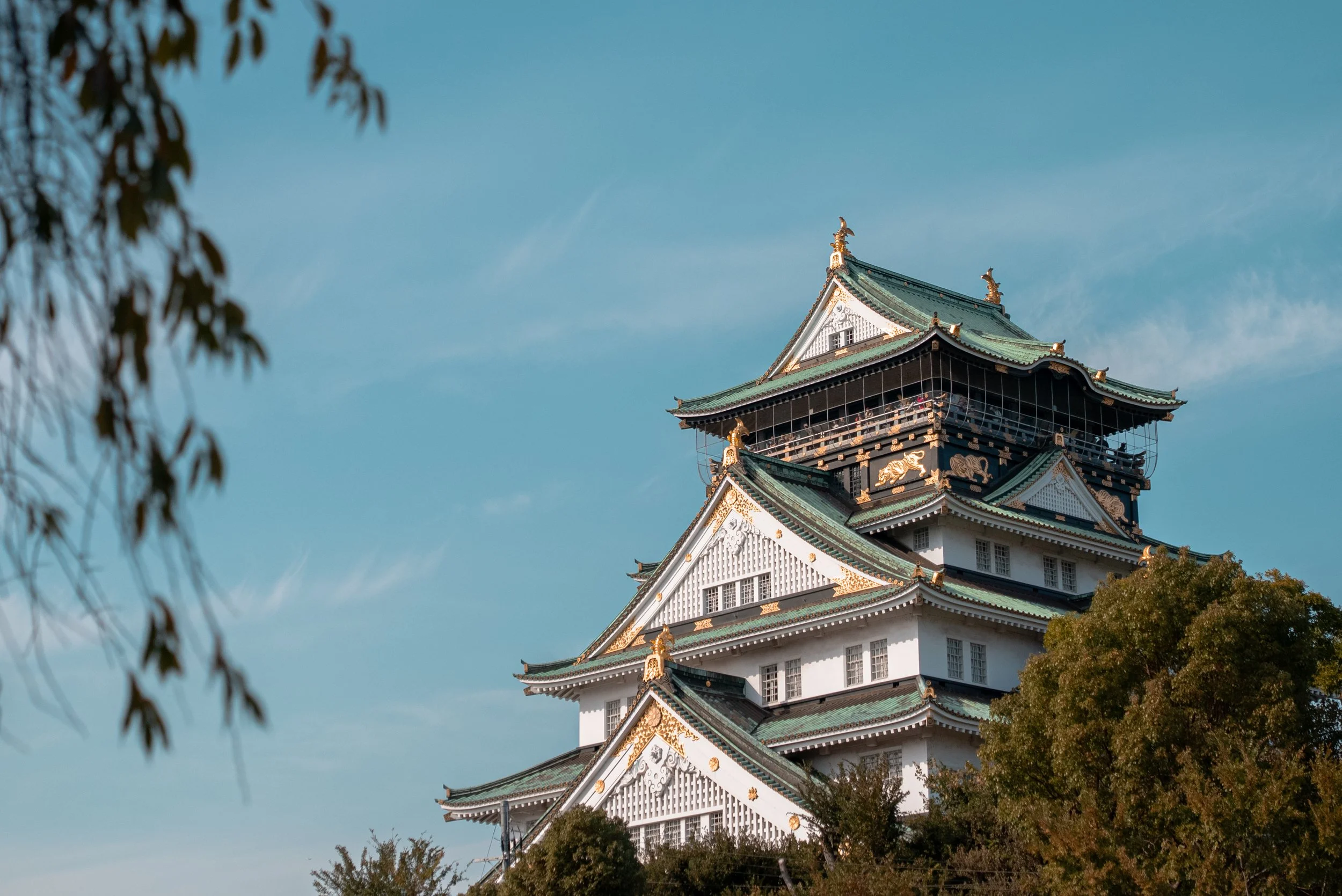 Château japonais traditionnel avec toits verts et ornements dorés, entouré d'arbres, sous un ciel bleu.