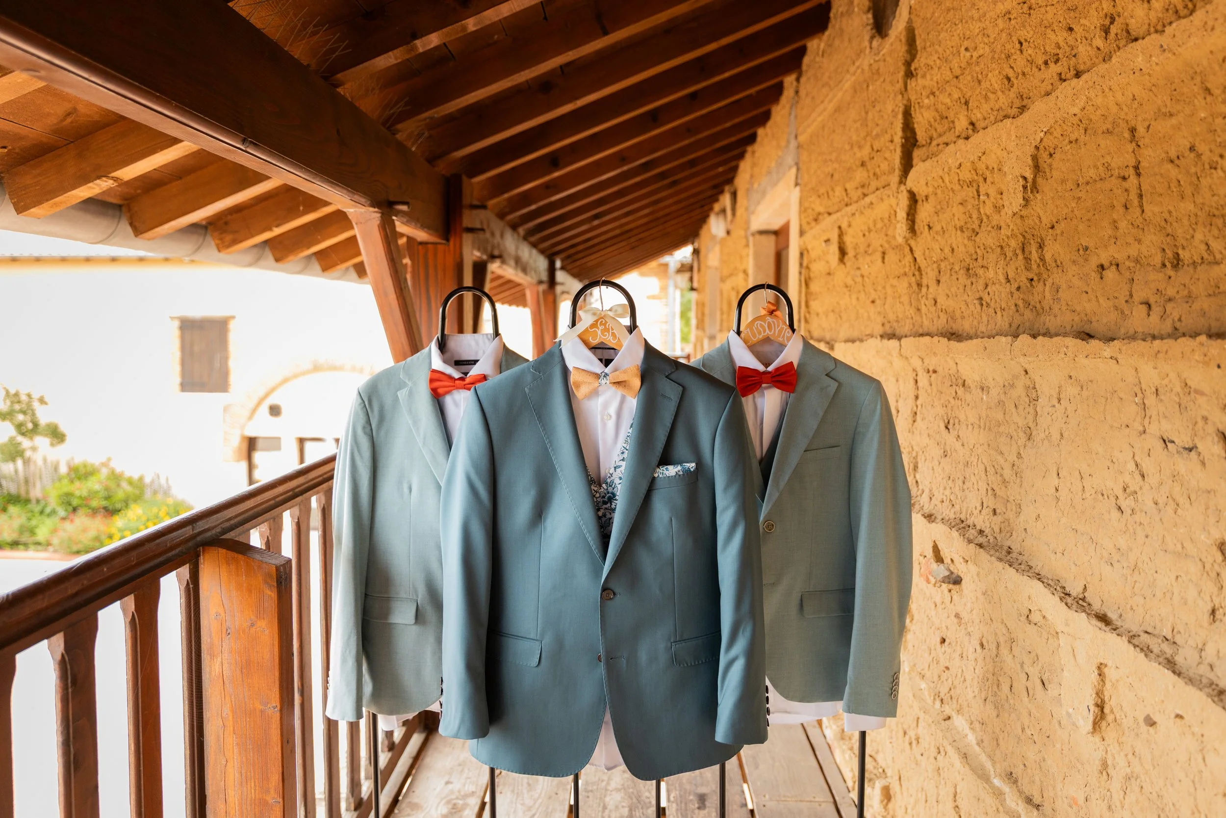 Trois costumes bleus avec chemises blanches et nœuds papillons rouges ou beige suspendus sur des cintres sur un balcon en bois, avec un mur en pierre jaune en arrière-plan.