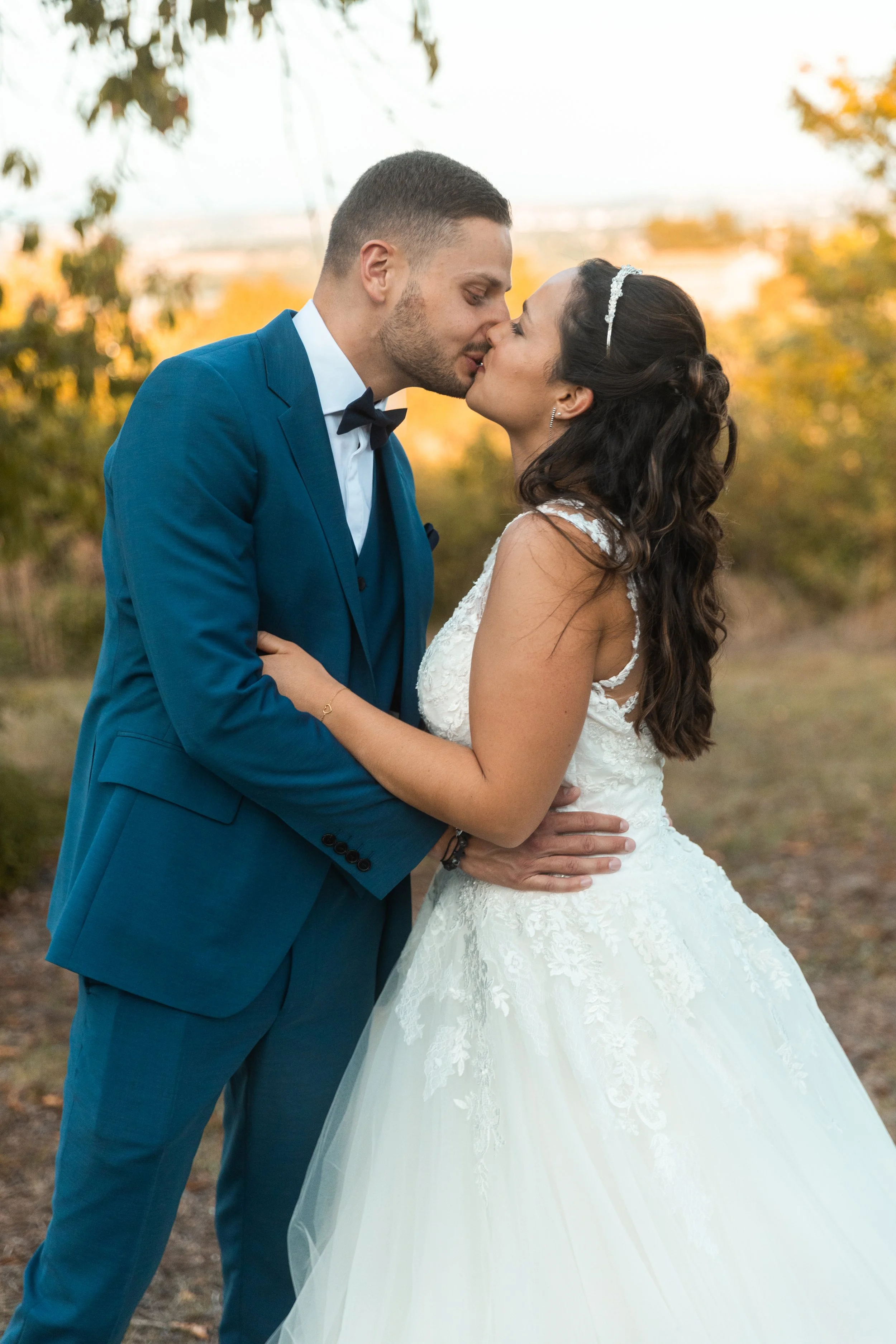 Un couple en mariée et marié s'embrassant lors d'une séance photo en plein air, avec des arbres aux feuilles d'automne en arrière-plan.