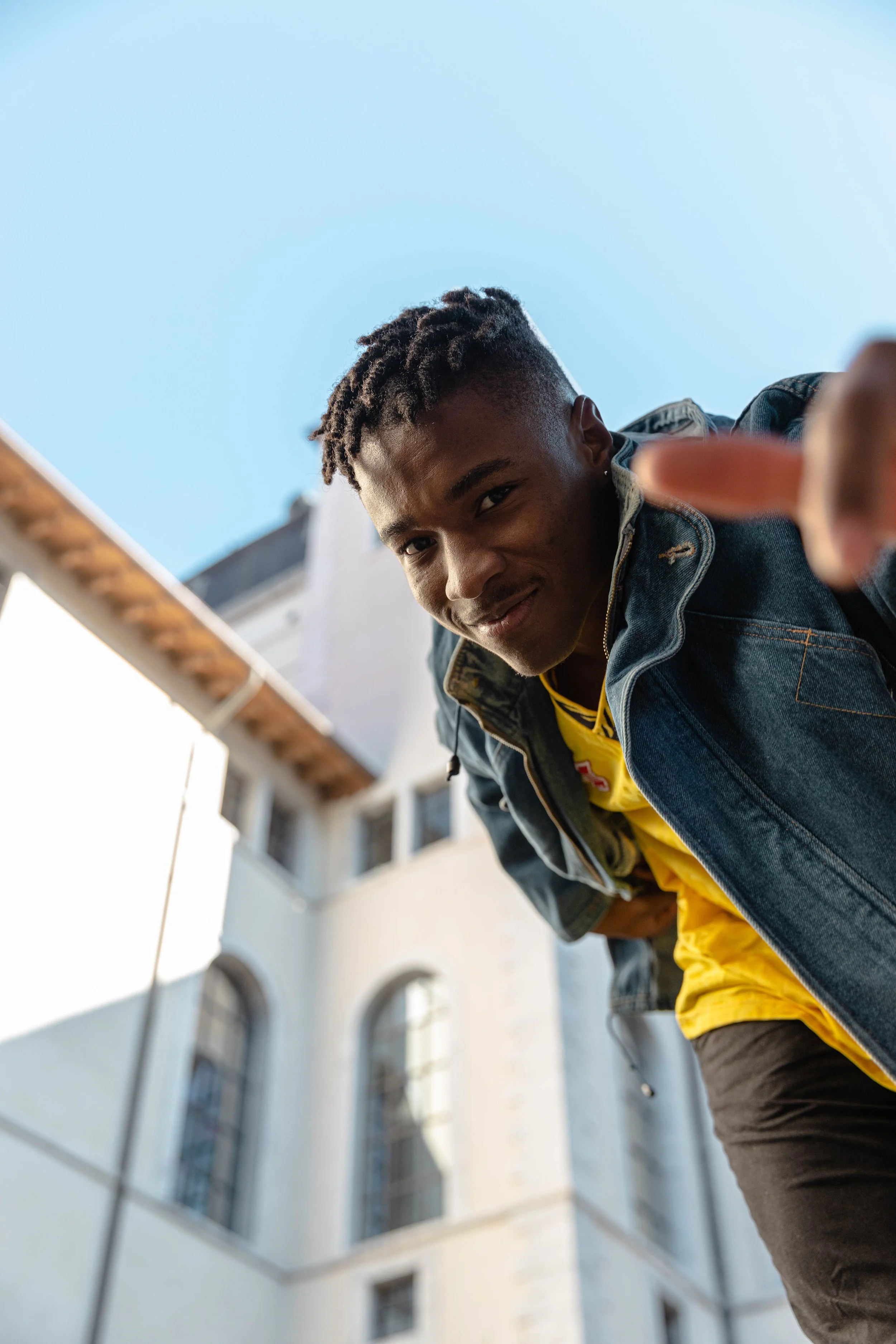 Jeune homme souriant, de race noire, regardant vers la caméra, en plein air, devant un bâtiment historique, portant une veste en jean et un t-shirt jaune.