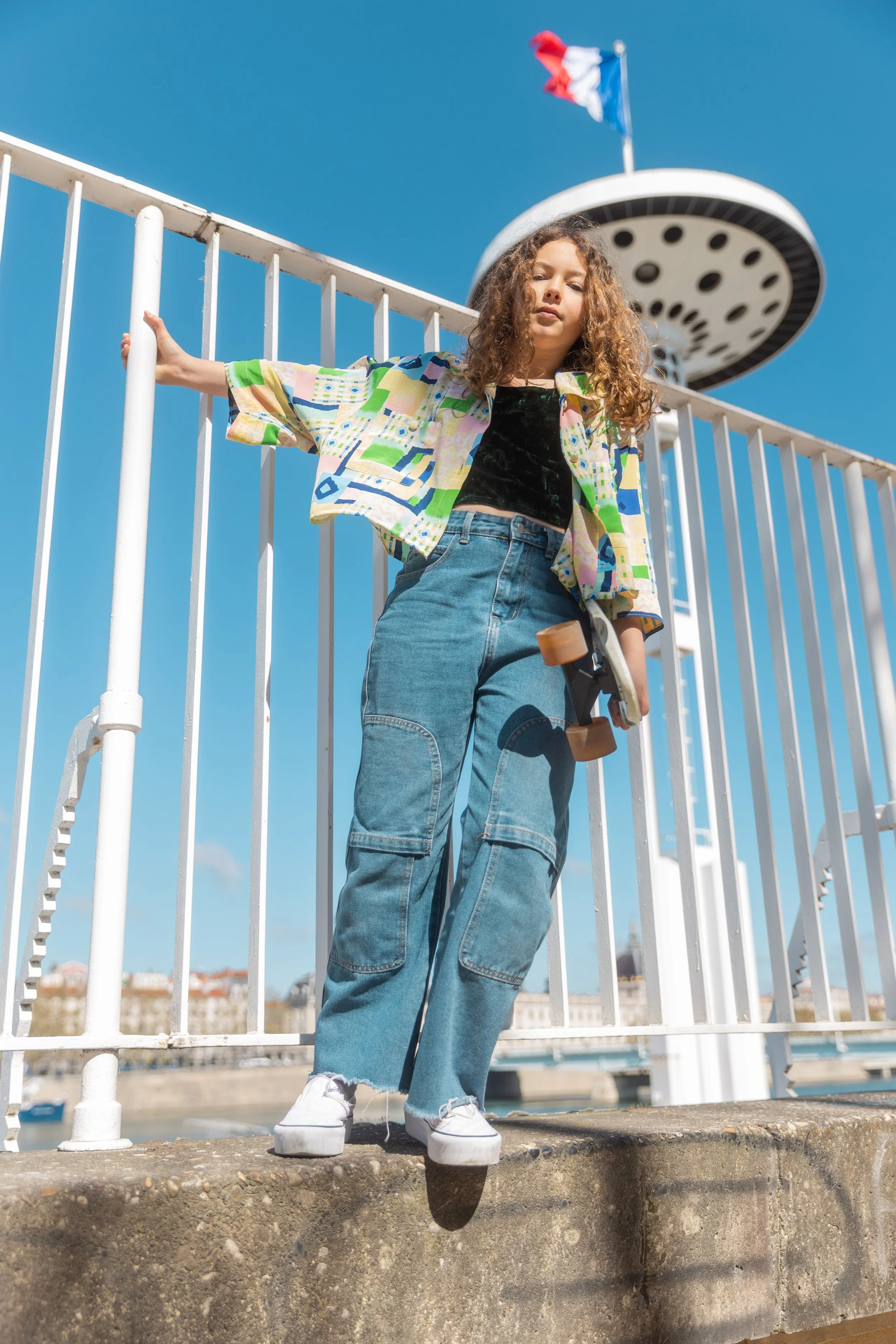 Jeune fille avec un skateboard, portant un chemisier coloré, un haut noir et un jean, debout devant une clôture en fer blanc avec un ciel bleu et une tour d'observation en arrière-plan.