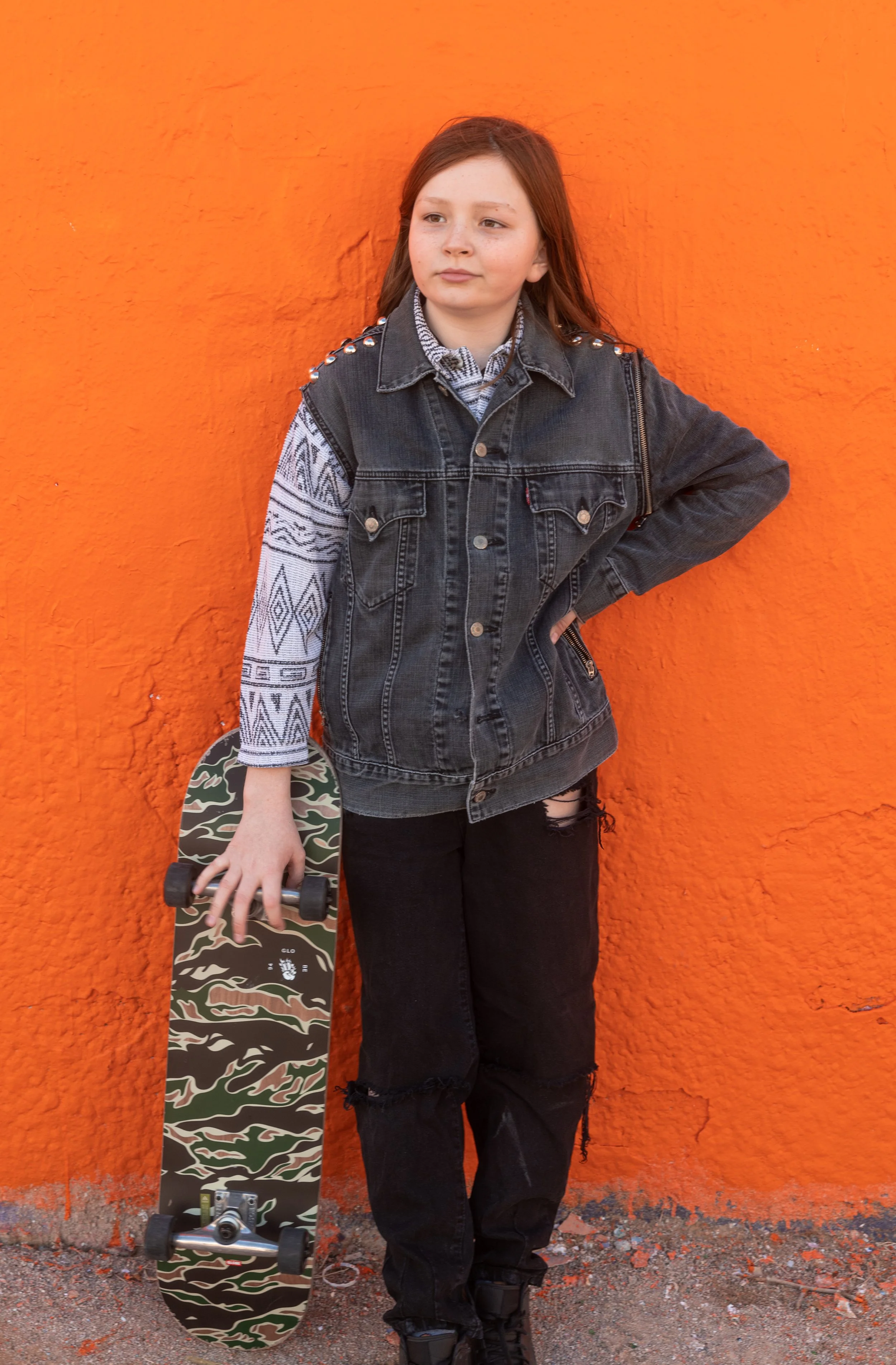 Une jeune fille avec des cheveux roux, portant une veste en denim noire et un pantalon noir déchiré, tenant un skateboard à motifs camouflage, se tient contre un mur orange.