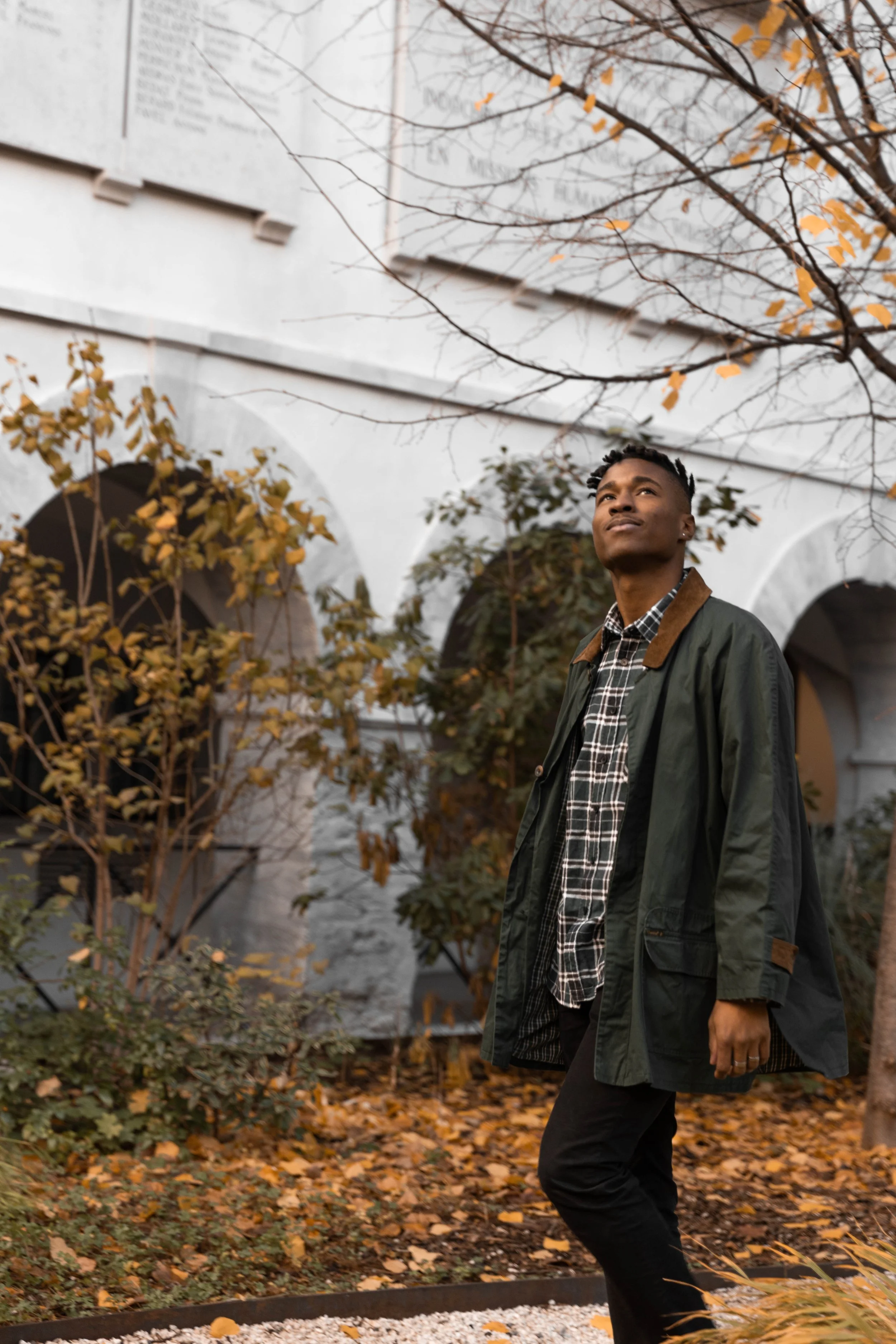 Un homme debout dans un jardin automnal, portant une veste verte et une chemise à carreaux, avec des feuilles d'automne tombées autour, sous un arbre aux branches sans beaucoup de feuilles.