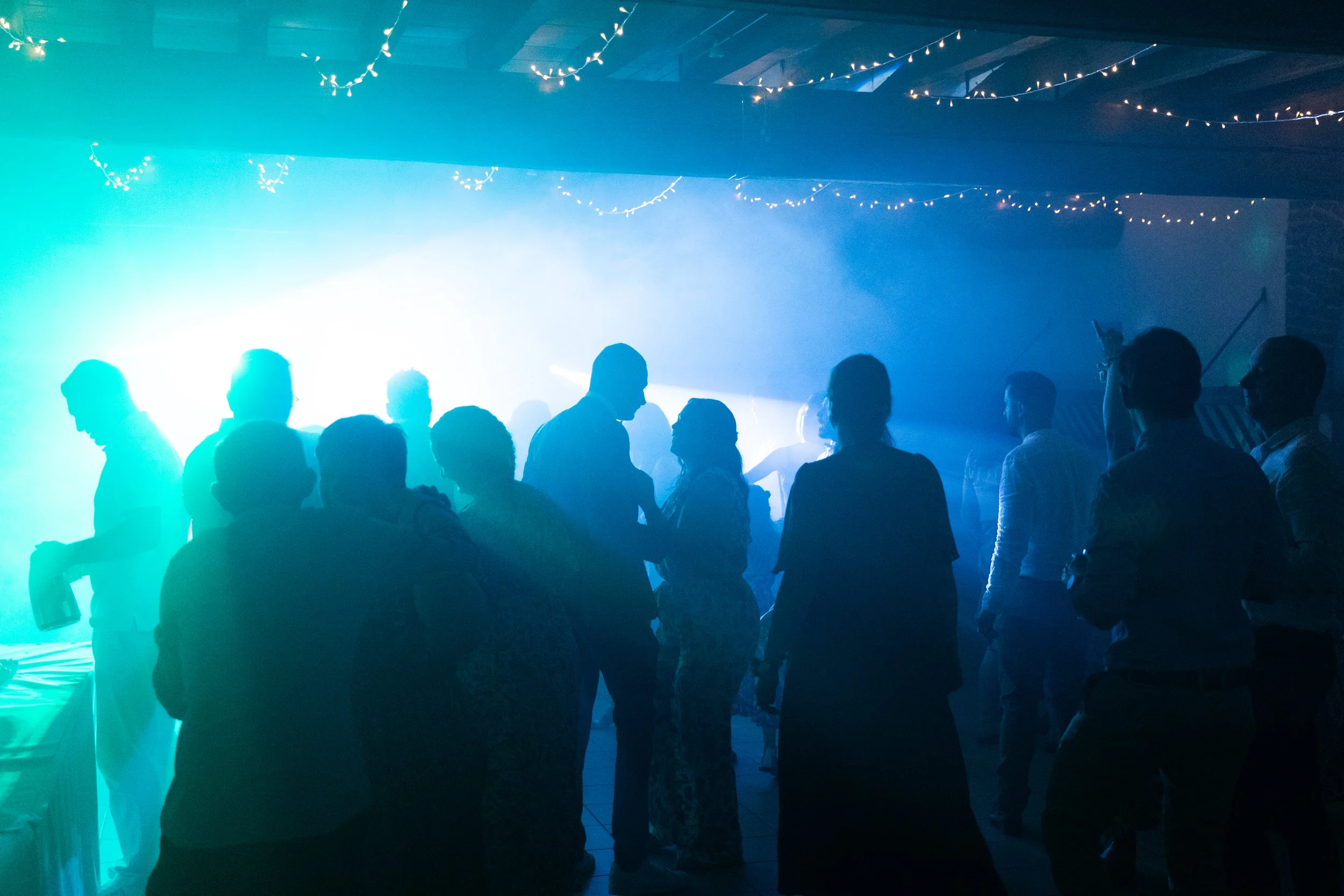 Des silhouettes de personnes dansants lors d'une fête ou soirée, sous un éclairage coloré avec des lumières suspendues décoratives.
