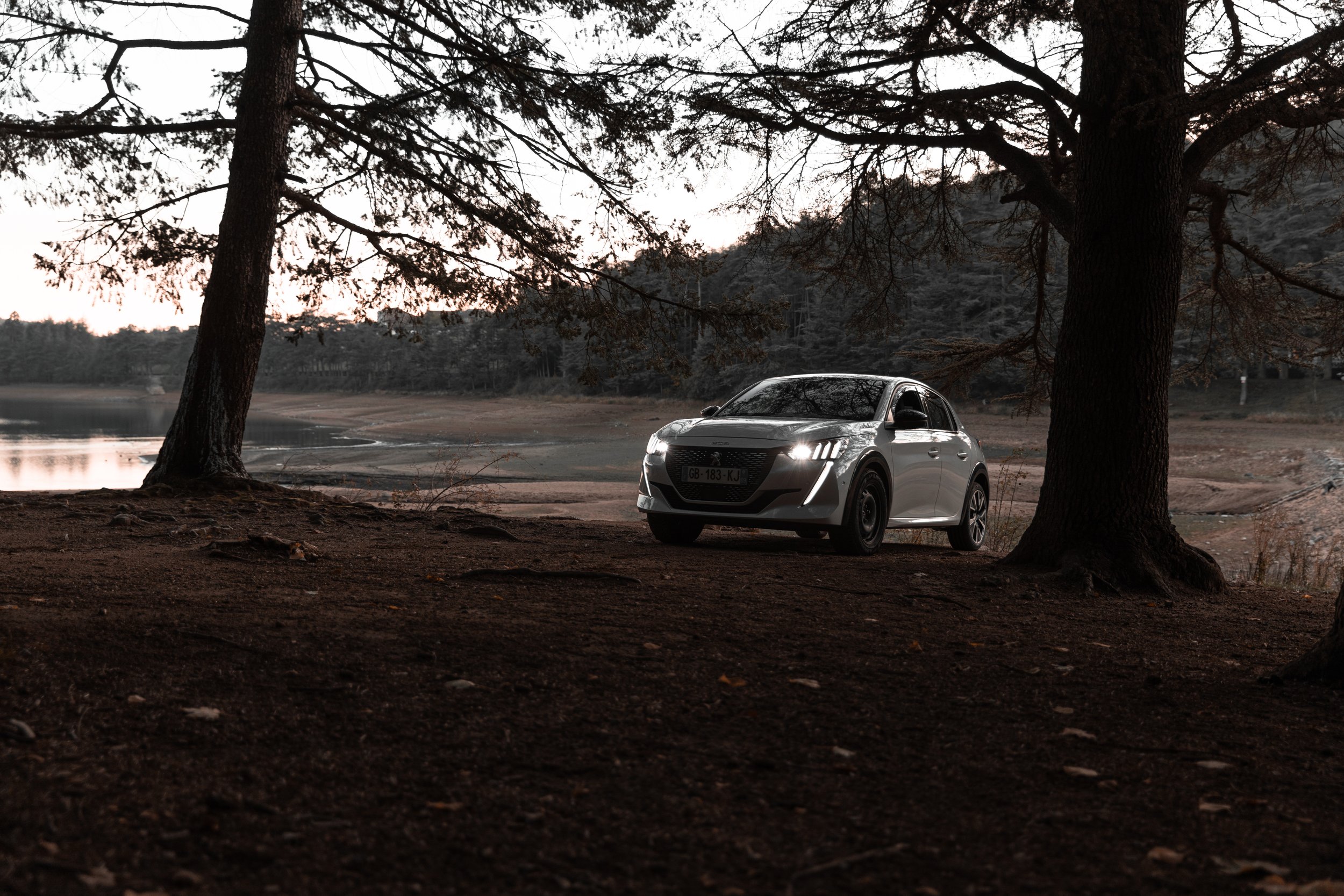 Une voiture grise stationnée sous un arbre au bord d'un lac, avec un paysage naturel au coucher du soleil ou au crépuscule.