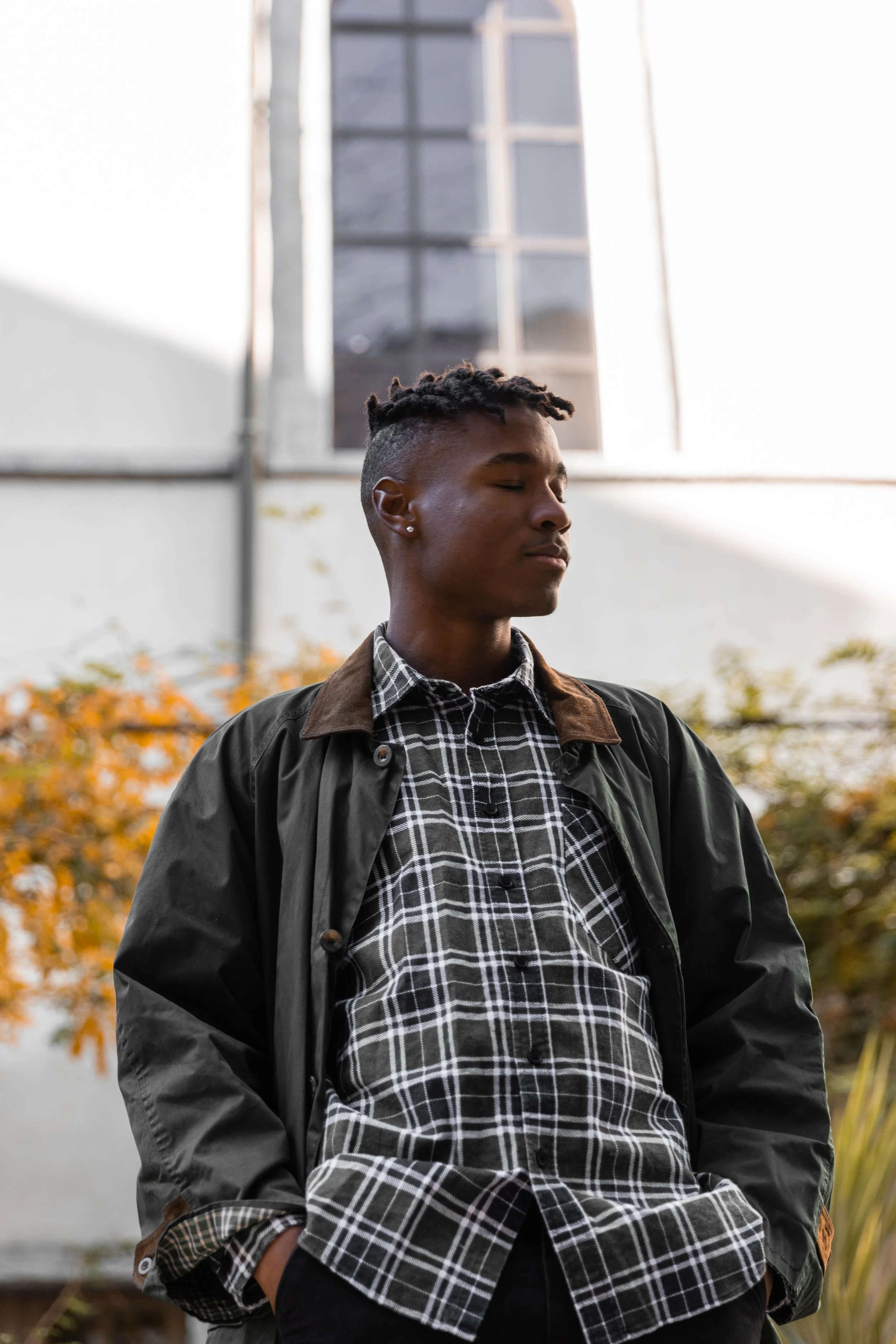 Jeune homme avec coiffure en torsades, portant une chemise à carreaux et une veste noire, se tenant dehors avec des feuilles d'automne en arrière-plan.