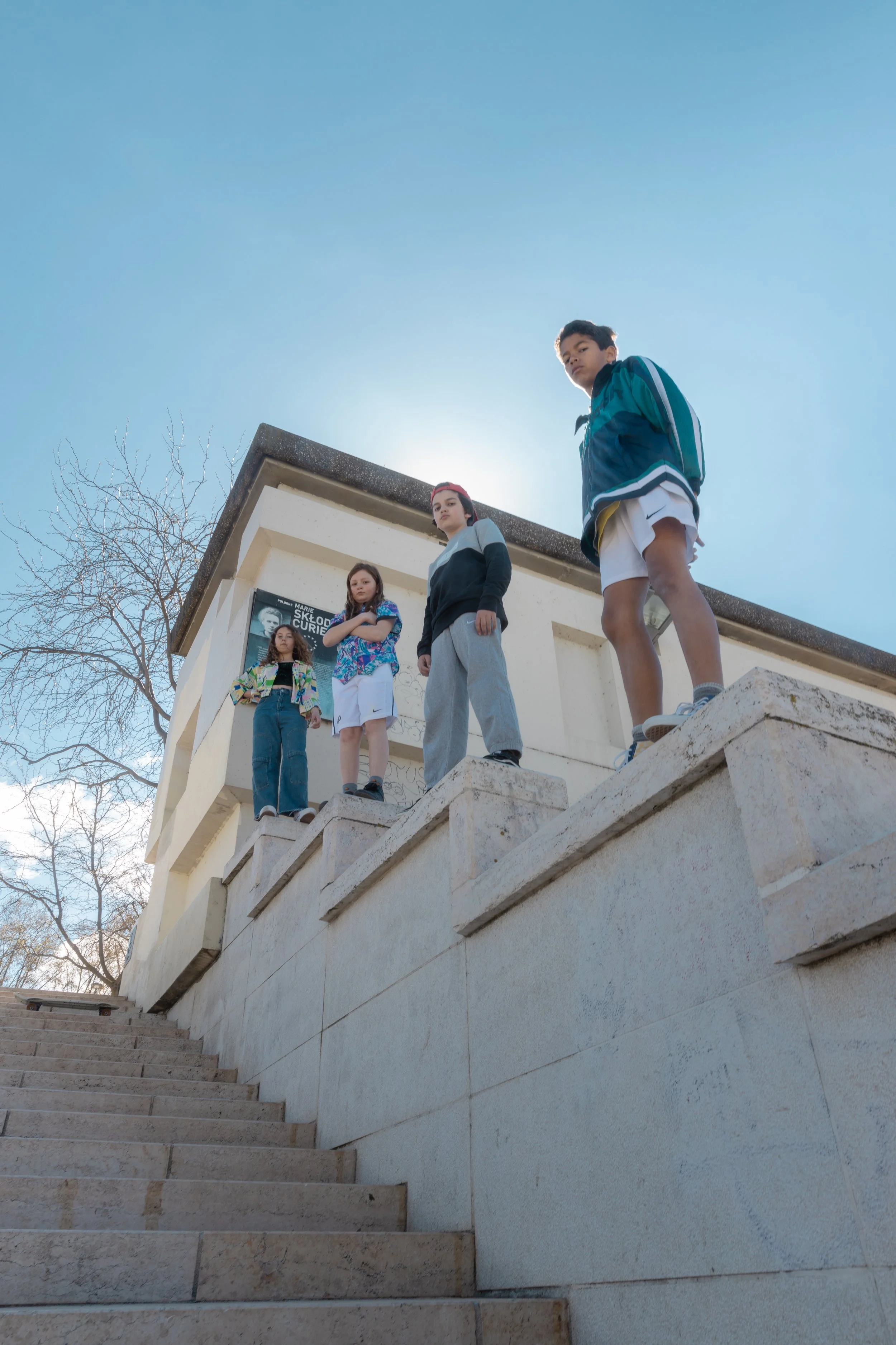 Quatre adolescents se tiennent sur une balustrade en pierre en haut d'un escalier extérieur, avec un bâtiment en arrière-plan et un ciel bleu clair.