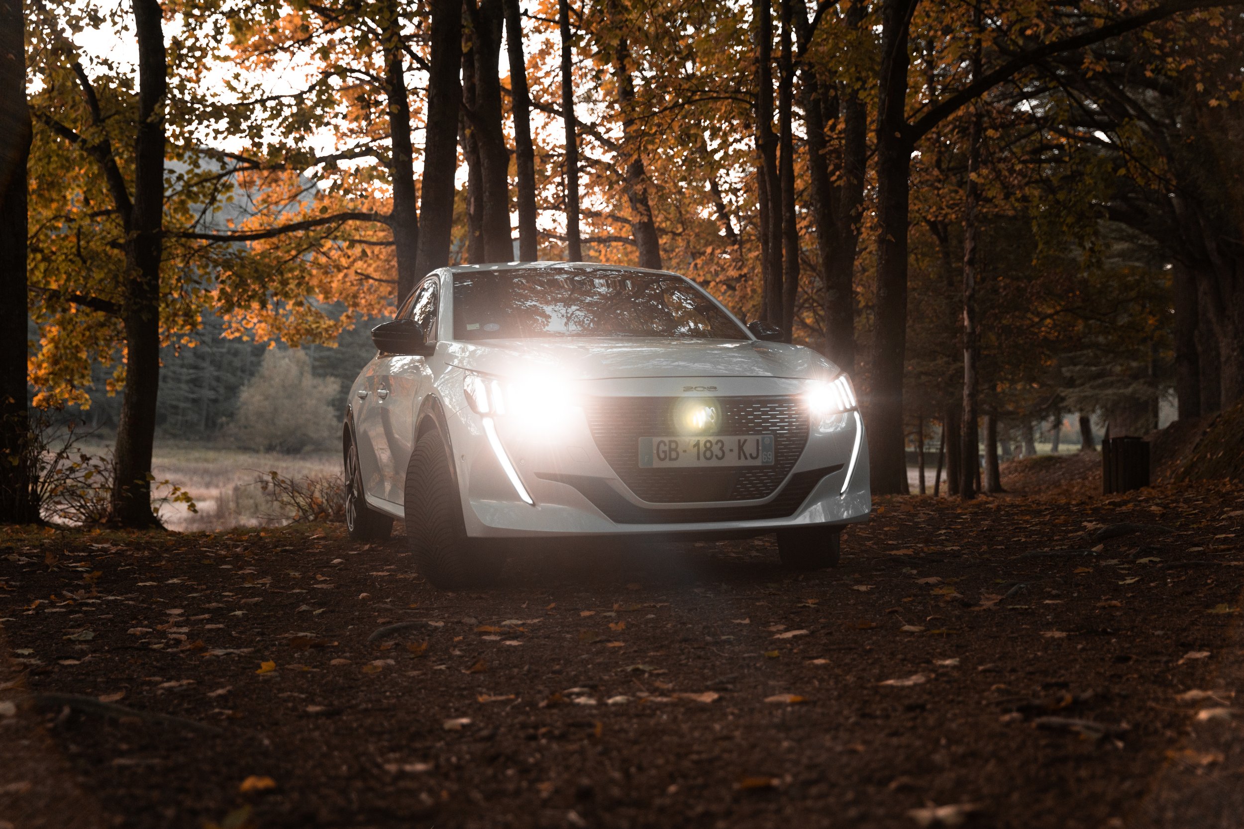 Une voiture blanche est garée sur un chemin forestier en automne, avec des feuilles tombées, et ses phares allumés. Il y a des arbres aux feuillages orange et vert en arrière-plan.