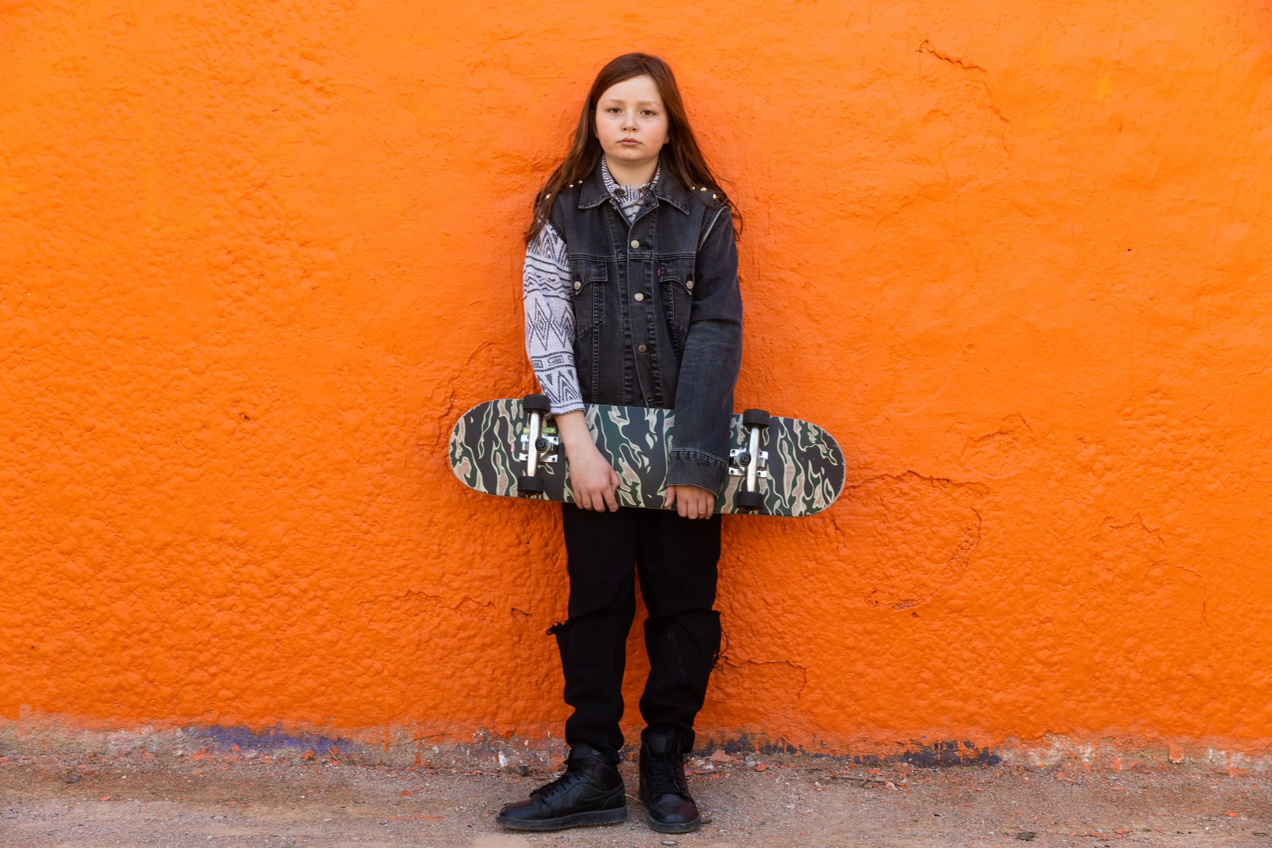 Une jeune fille avec des cheveux bruns, portant un jean sans manches gris et une veste en jean noire, tenant une planche à roulettes avec un motif camouflage, debout contre un mur orange.