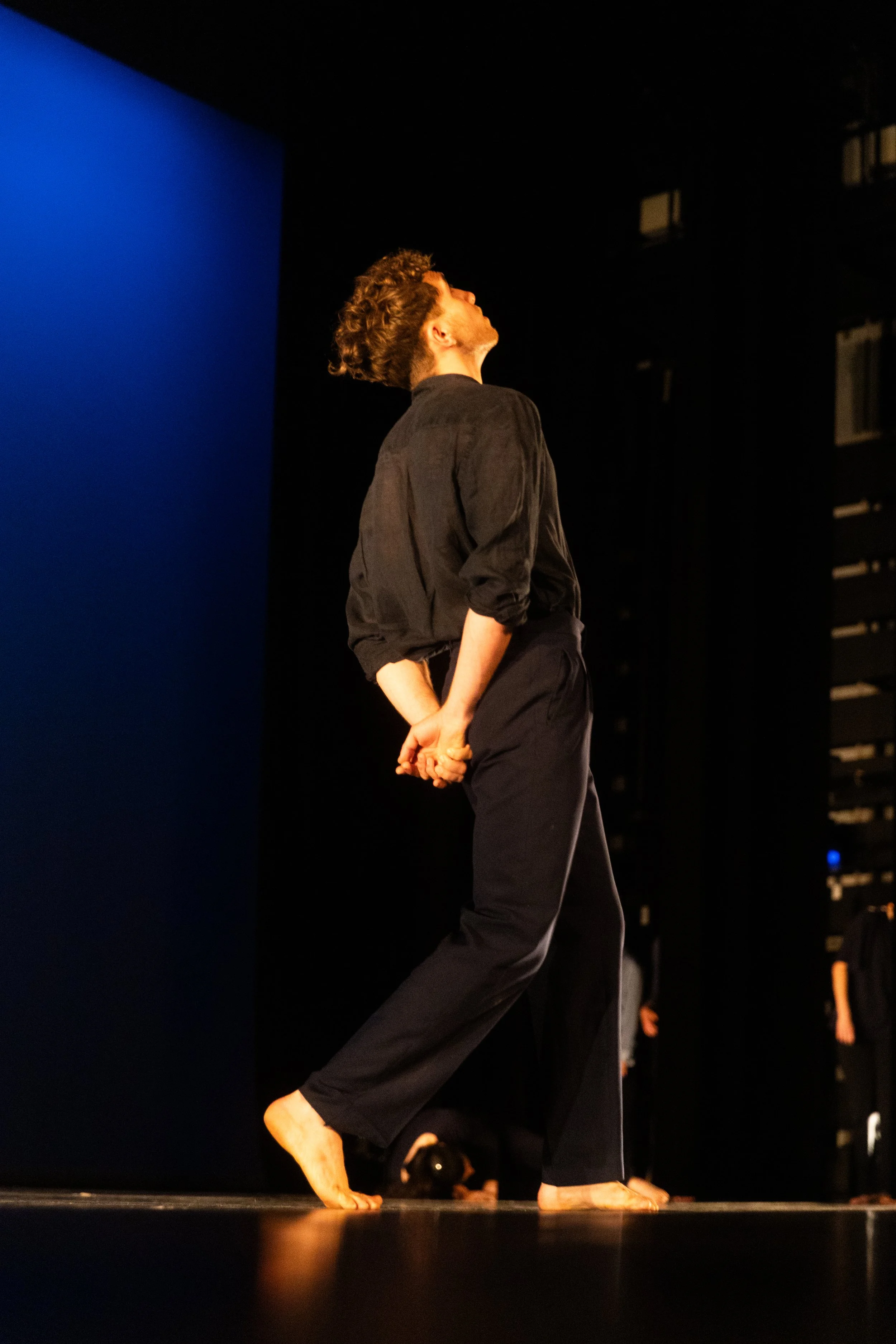 Une personne danse nue sur scène, tenant ses mains derrière le dos, avec un fond noir et un mur bleu. Elle porte une chemise noire et un pantalon noir.