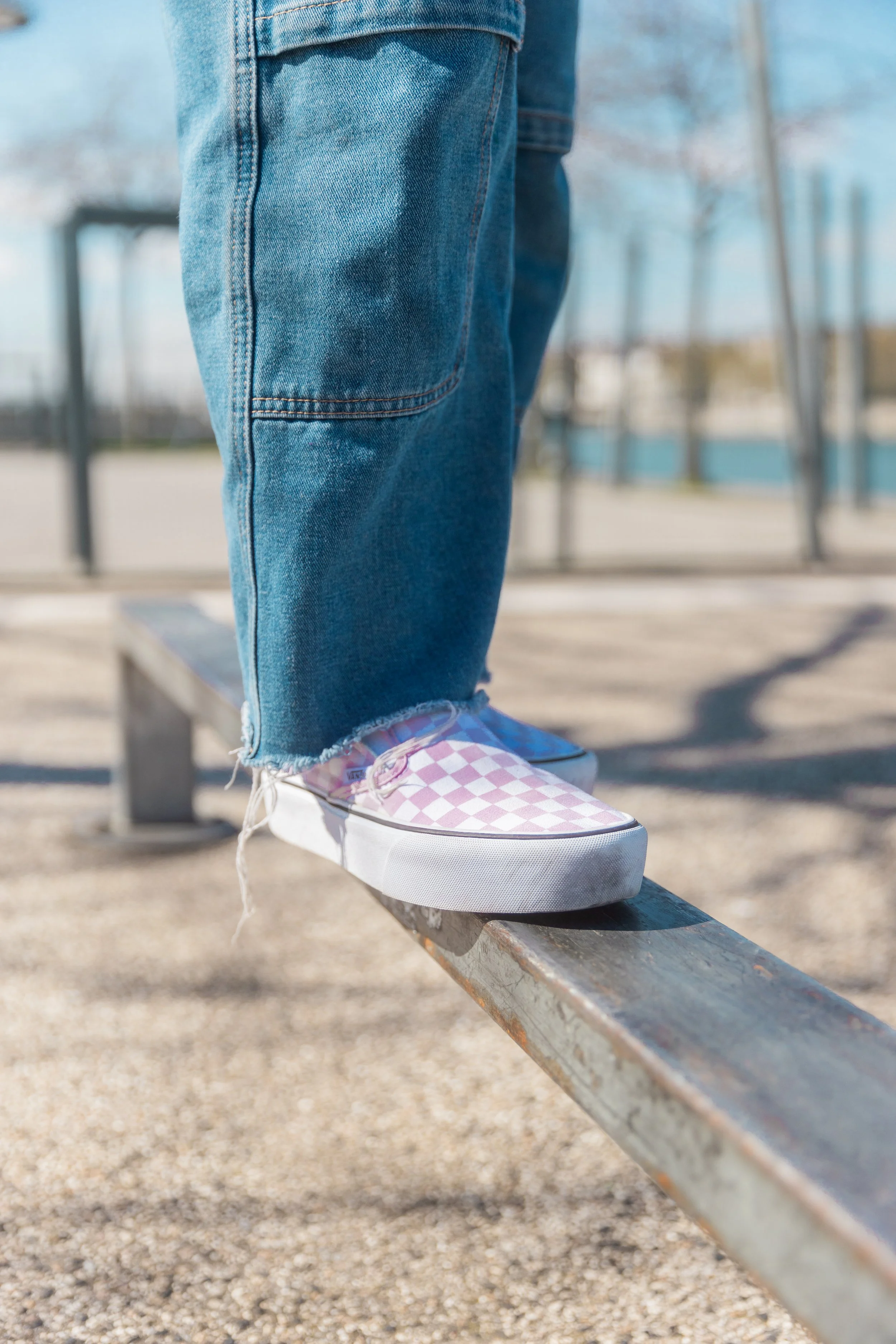 Person qui équilibre sur une barre métallique, portant des chaussures à motif vichy rose et blanc, et un jean déchiré à la cheville, dans un parc.