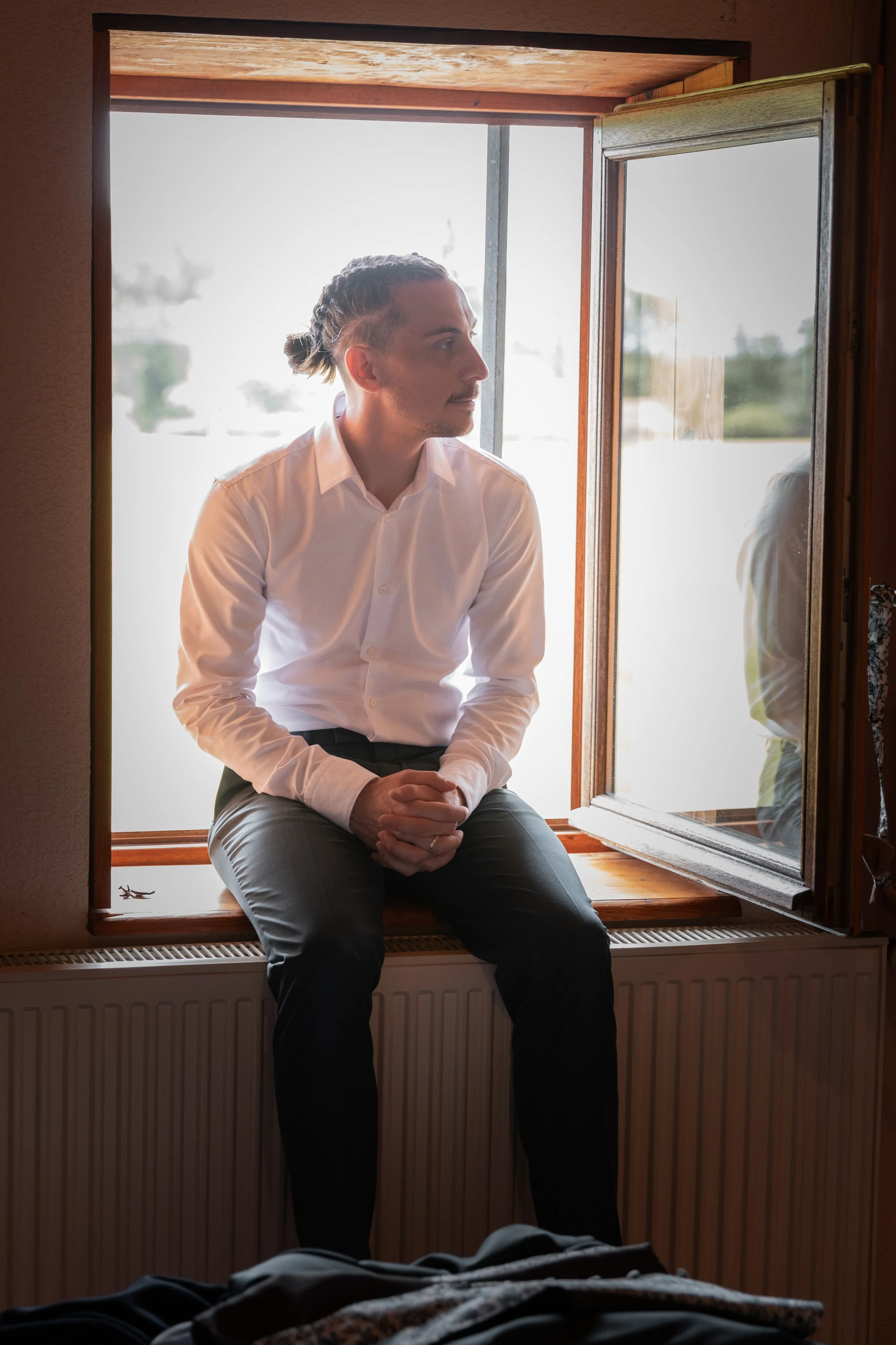 Un homme assis sur un rebord de fenêtre, portant une chemise blanche, regardant par la fenêtre ouverte, dans une pièce lumineuse.