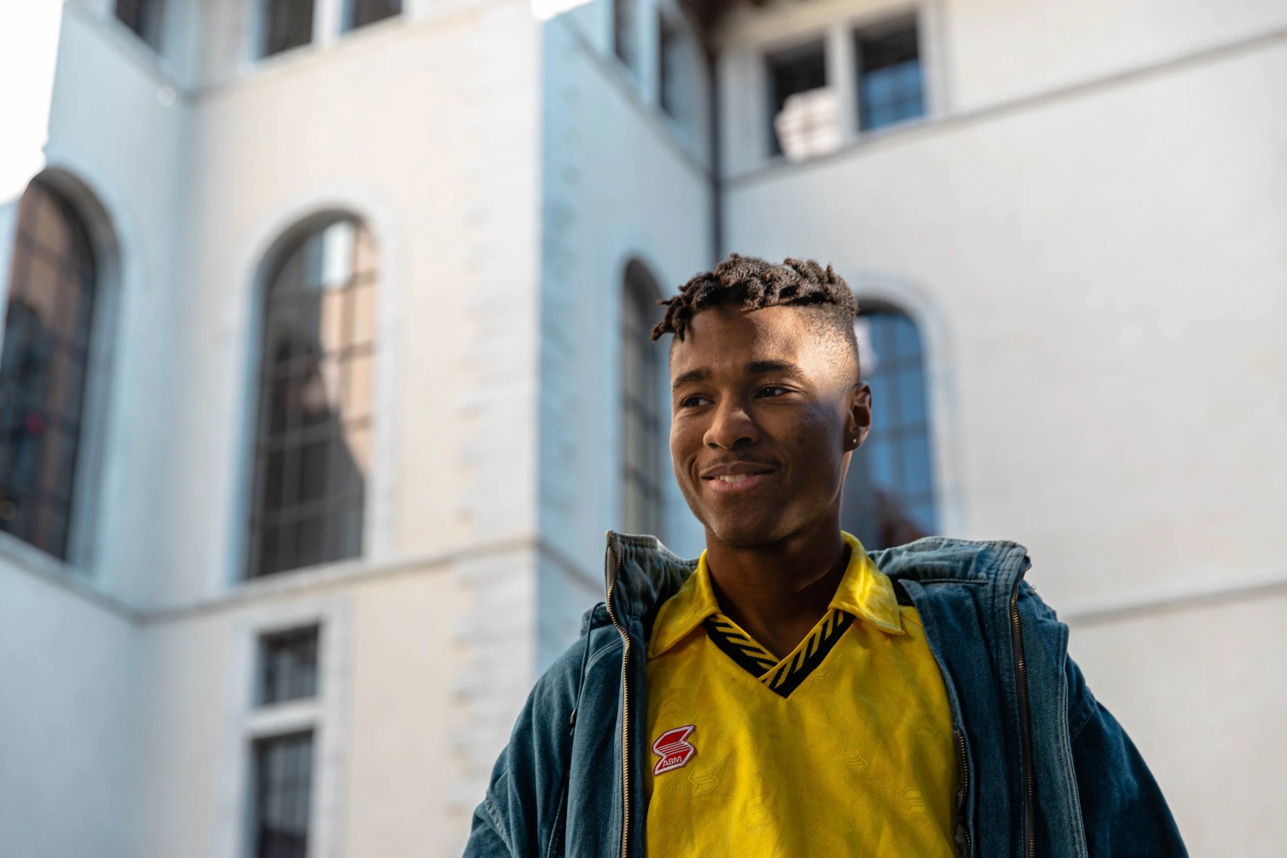 Un jeune homme souriant portant un hoodie en jean et une chemise jaune se tient devant un bâtiment blanc avec de grandes fenêtres.