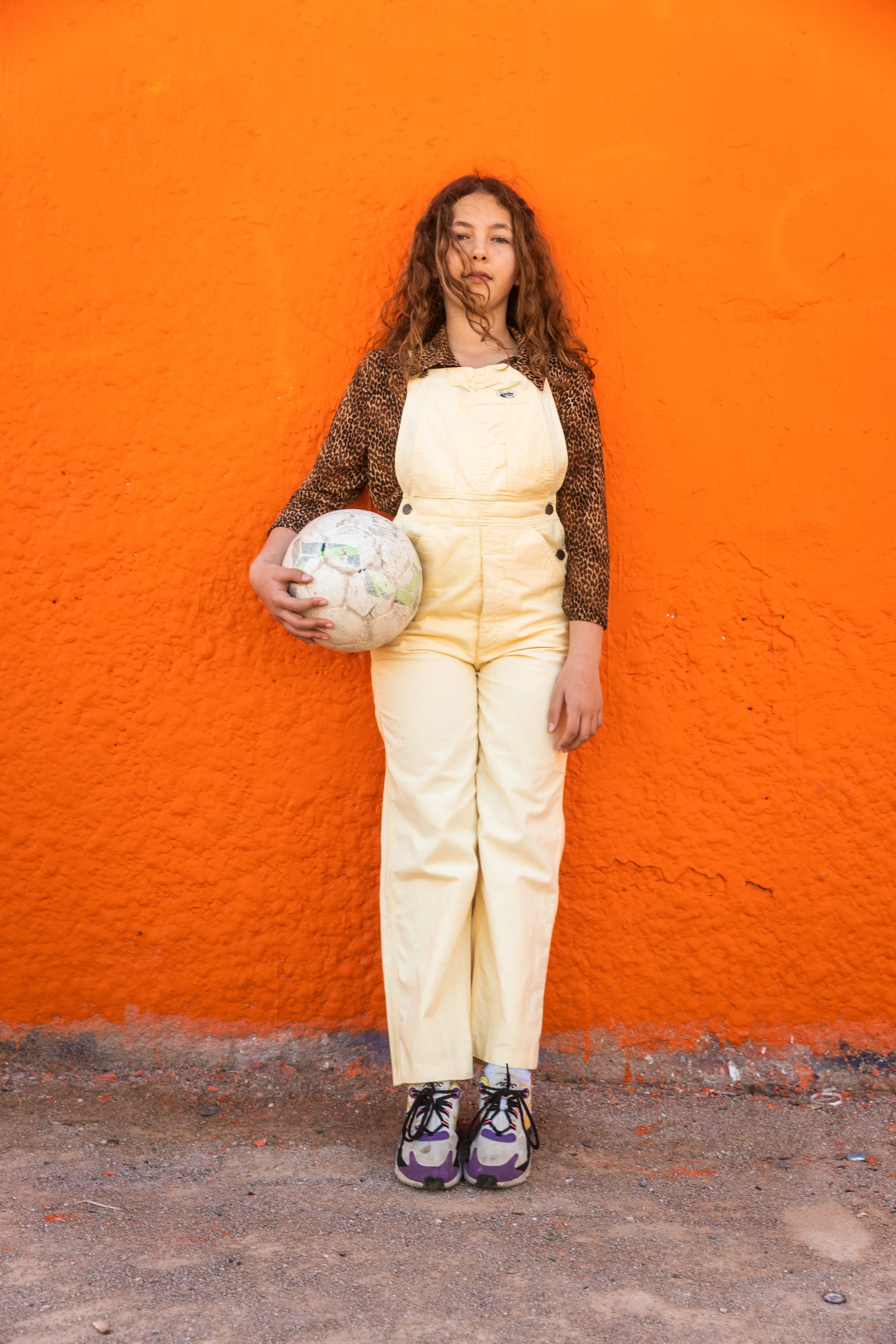 Jeune fille posant contre un mur orange, tenant un ballon de football usé, portant un vêtement à motif léopard sur la veste, une combinaison beige et des baskets colorées.