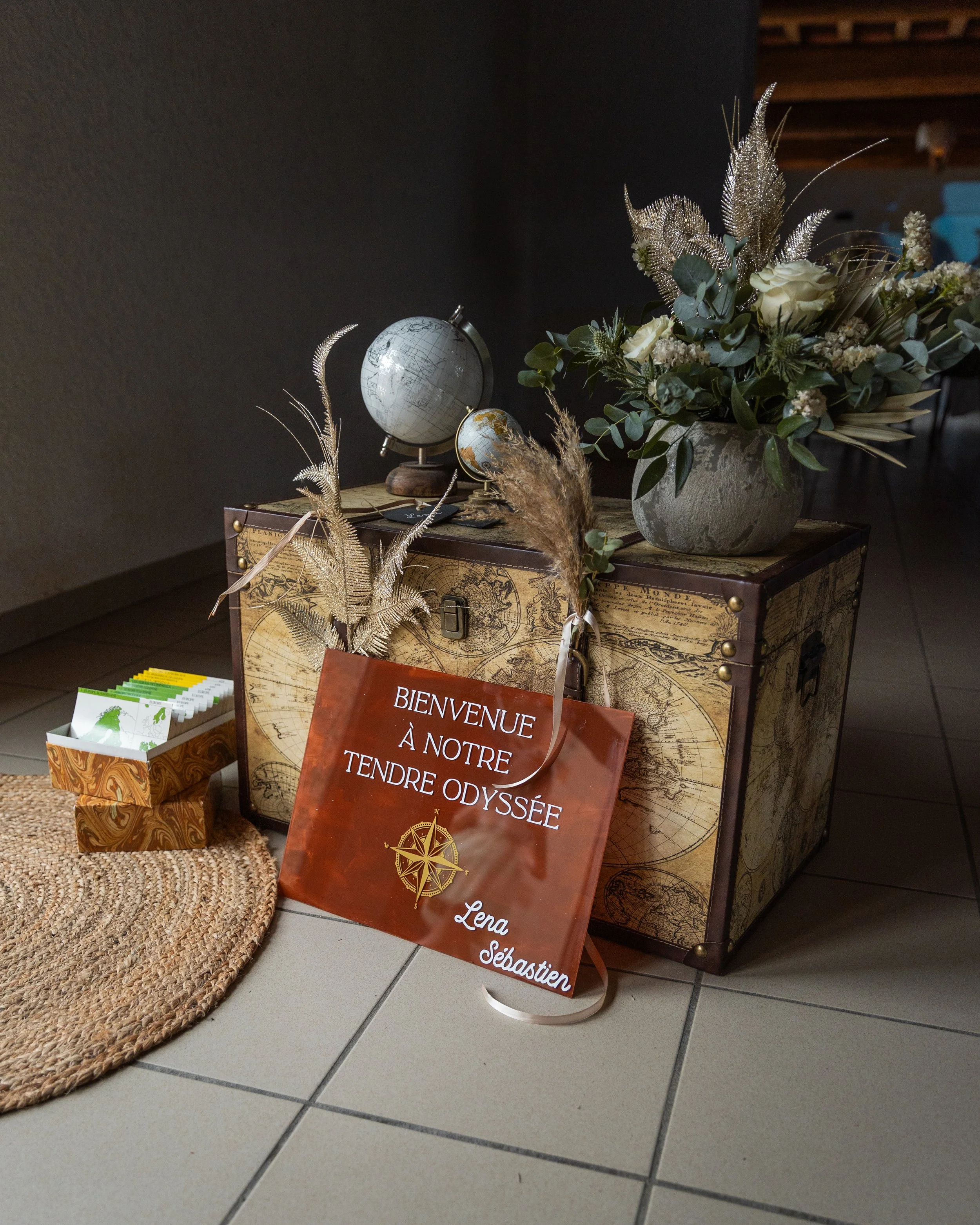 Une caisse en bois avec une décoration de globe terrestre et un bouquet de fleurs dans un vase, avec un panneau en plastique indiquant "Bienvenue à notre tendre odyssée" et des noms "Lena" et "Sébastien".