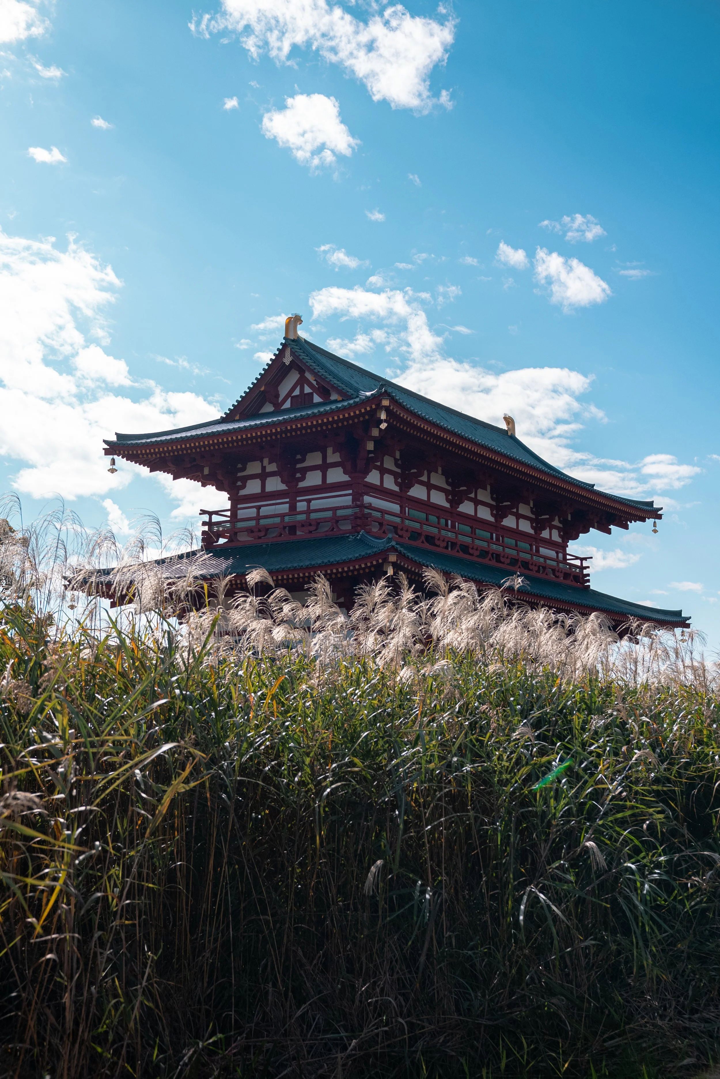 Une pagode traditionnelle japonaise avec un toit vert, entourée de hautes herbes, sous un ciel bleu avec quelques nuages.