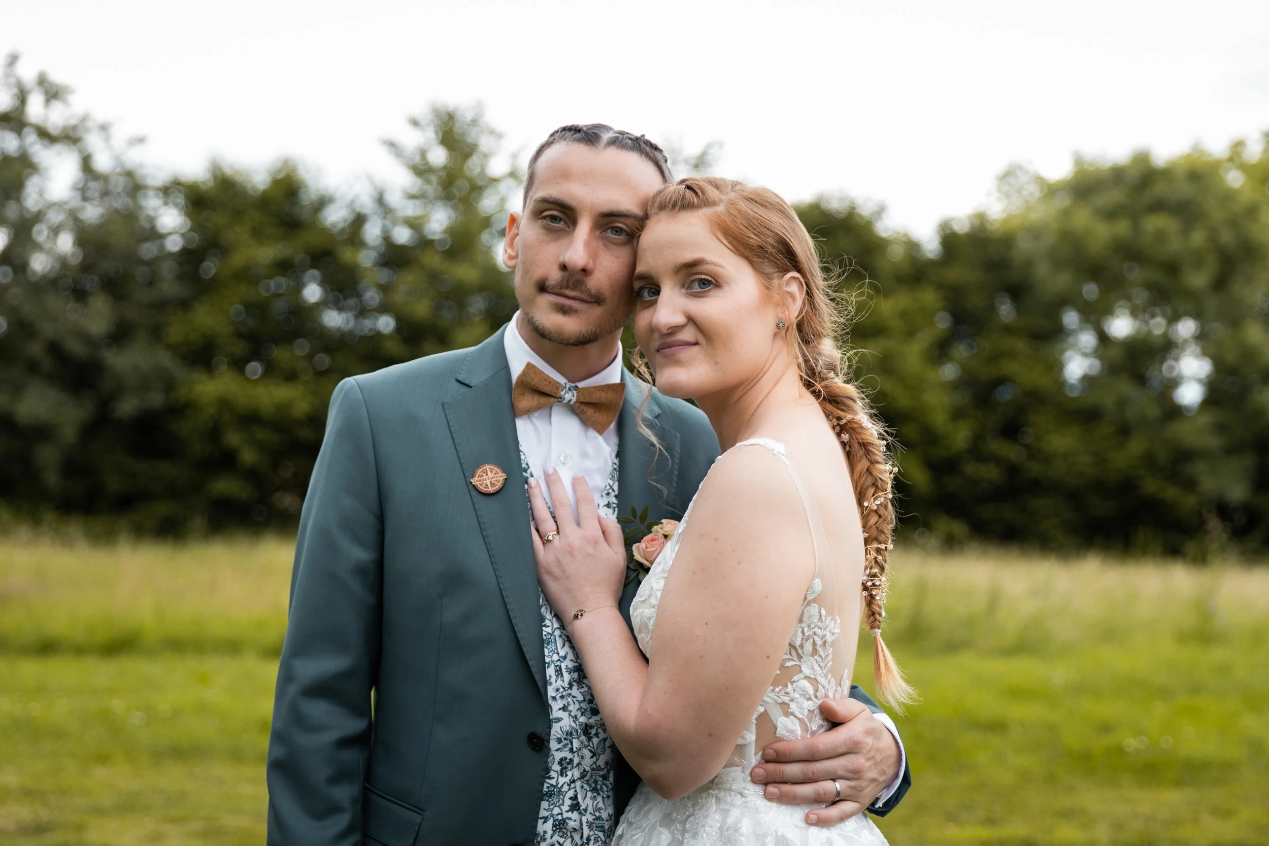 Un couple de mariage posant dans un champ vert, la femme porte une robe de mariée blanche et le homme un costume bleu avec un nœud papillon marron.