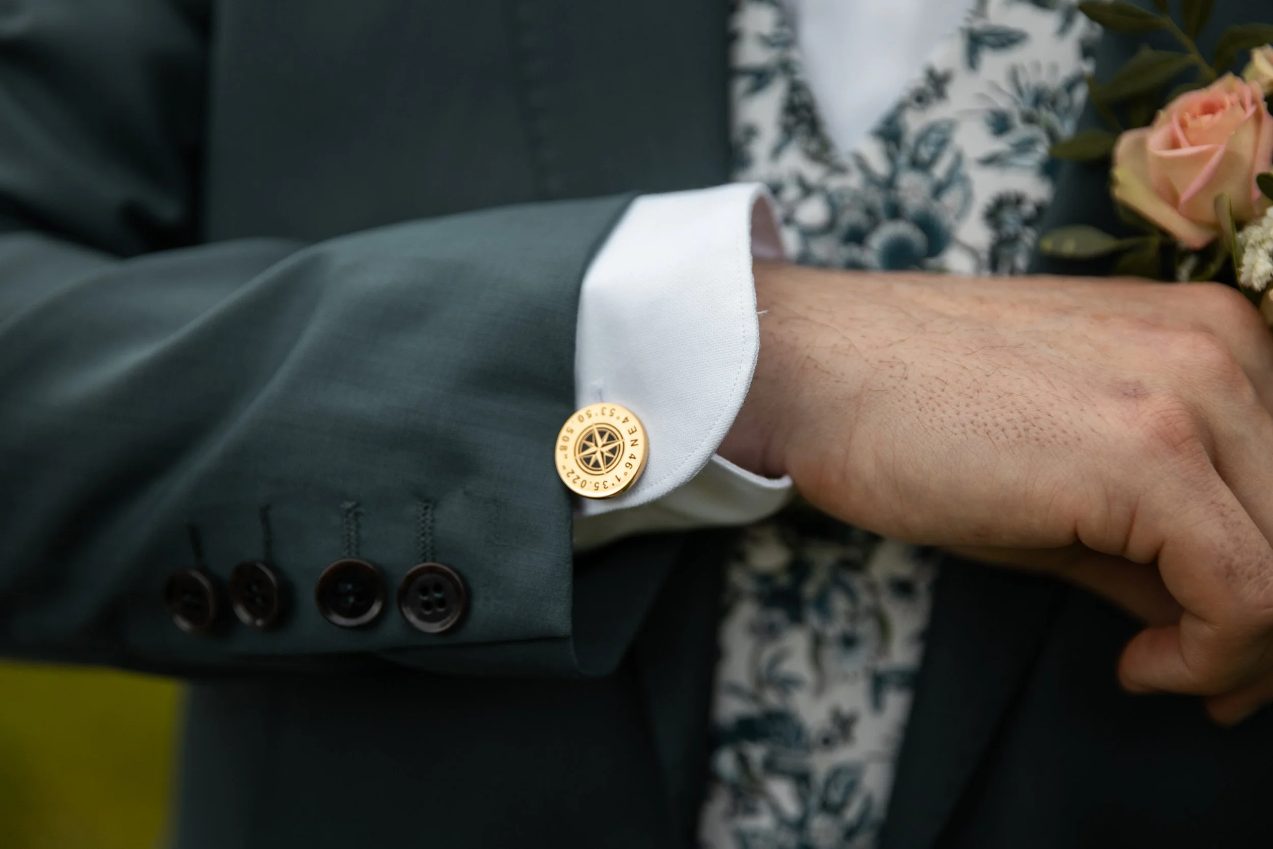 Poignet d'une personne en costume avec boutonnière en or, portant une chemise blanche aux manches longues, tenant un bouquet de fleurs.