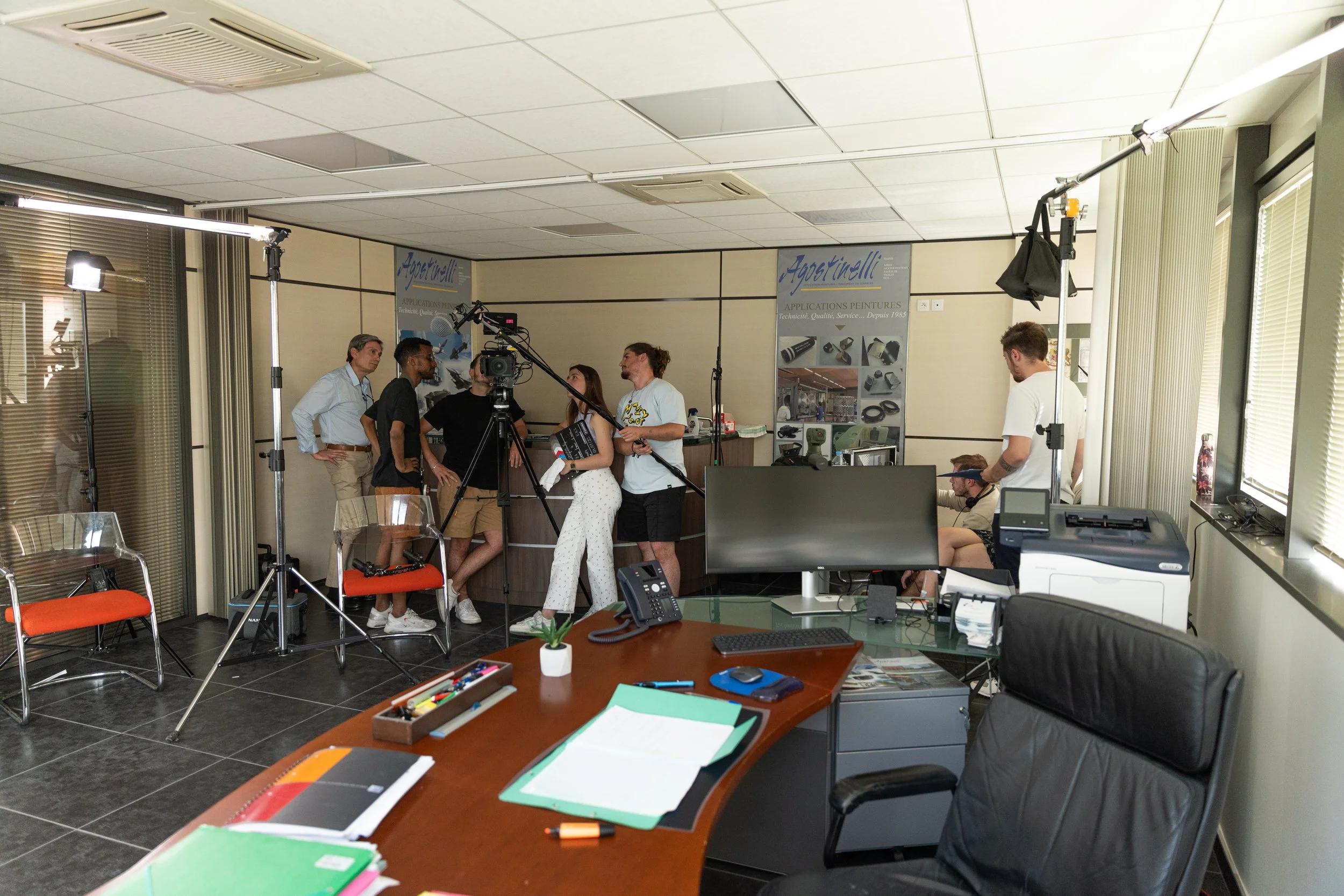 Une équipe de tournage prépare une interview ou une vidéo dans un bureau, avec plusieurs personnes, un caméraman, un panneau d'affichage, un bureau avec un ordinateur, un téléphone, des dossiers et une imprimante.