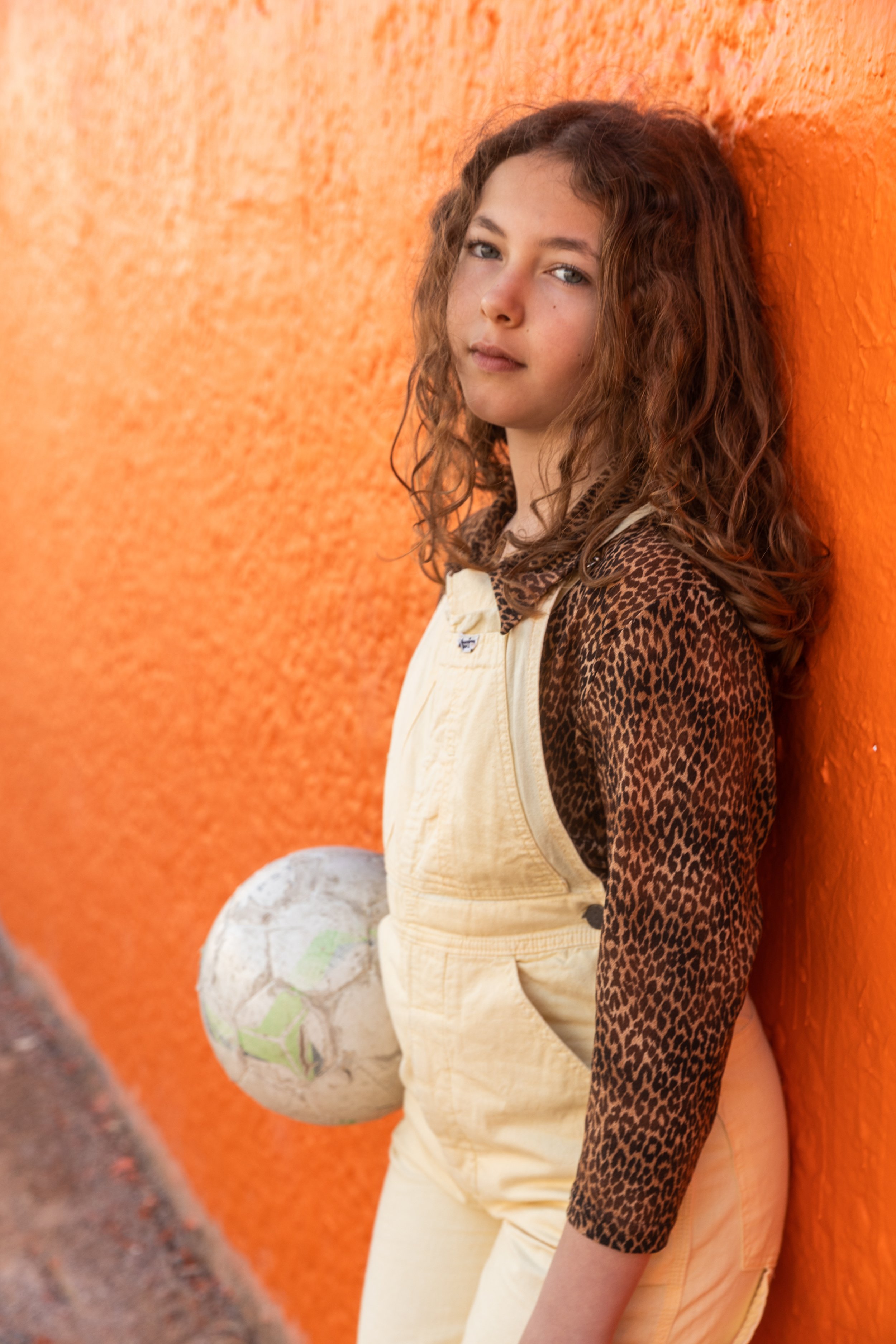 Jeune fille portant une veste léopard et des salopettes crème, tenant un ballon de football usé, appuyée contre un mur orange.
