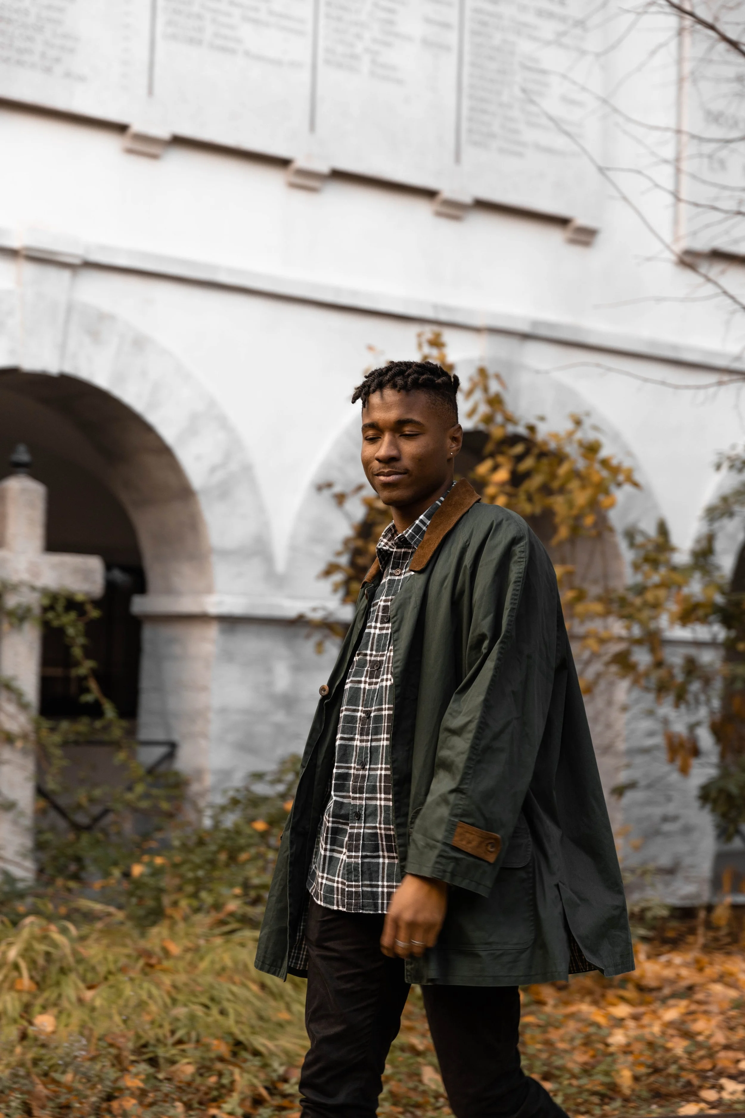 Un homme marchant dans un jardin avec des feuilles d'automne, portant une veste vert foncé et une chemise à carreaux, en arrière-plan un bâtiment blanc avec des arches.