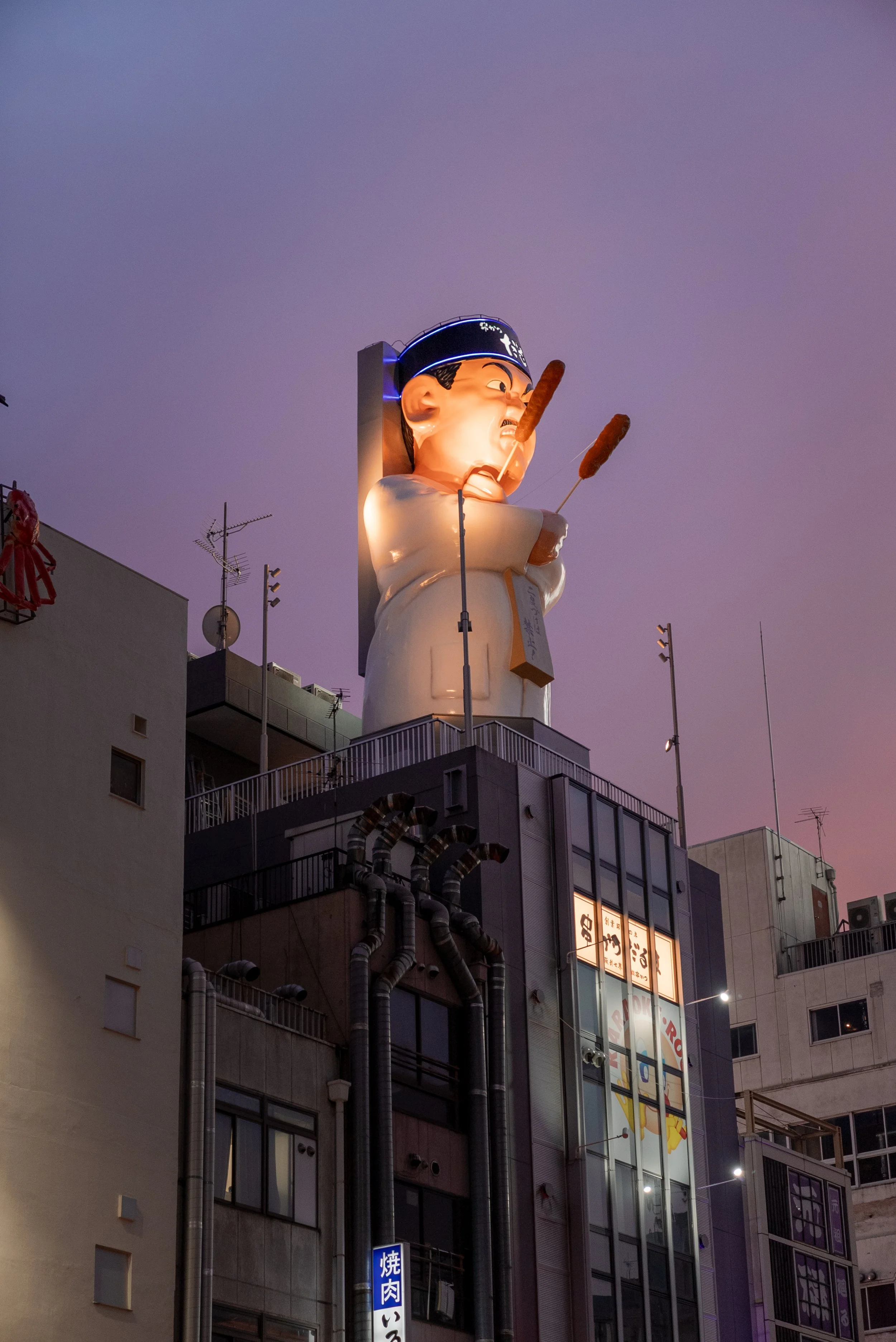 Une grande statue éclairée d'un chef japonais avec un chapeau en forme de toque, tenant deux brochettes, située sur le toit d'un bâtiment en ville, avec un ciel violet en arrière-plan.