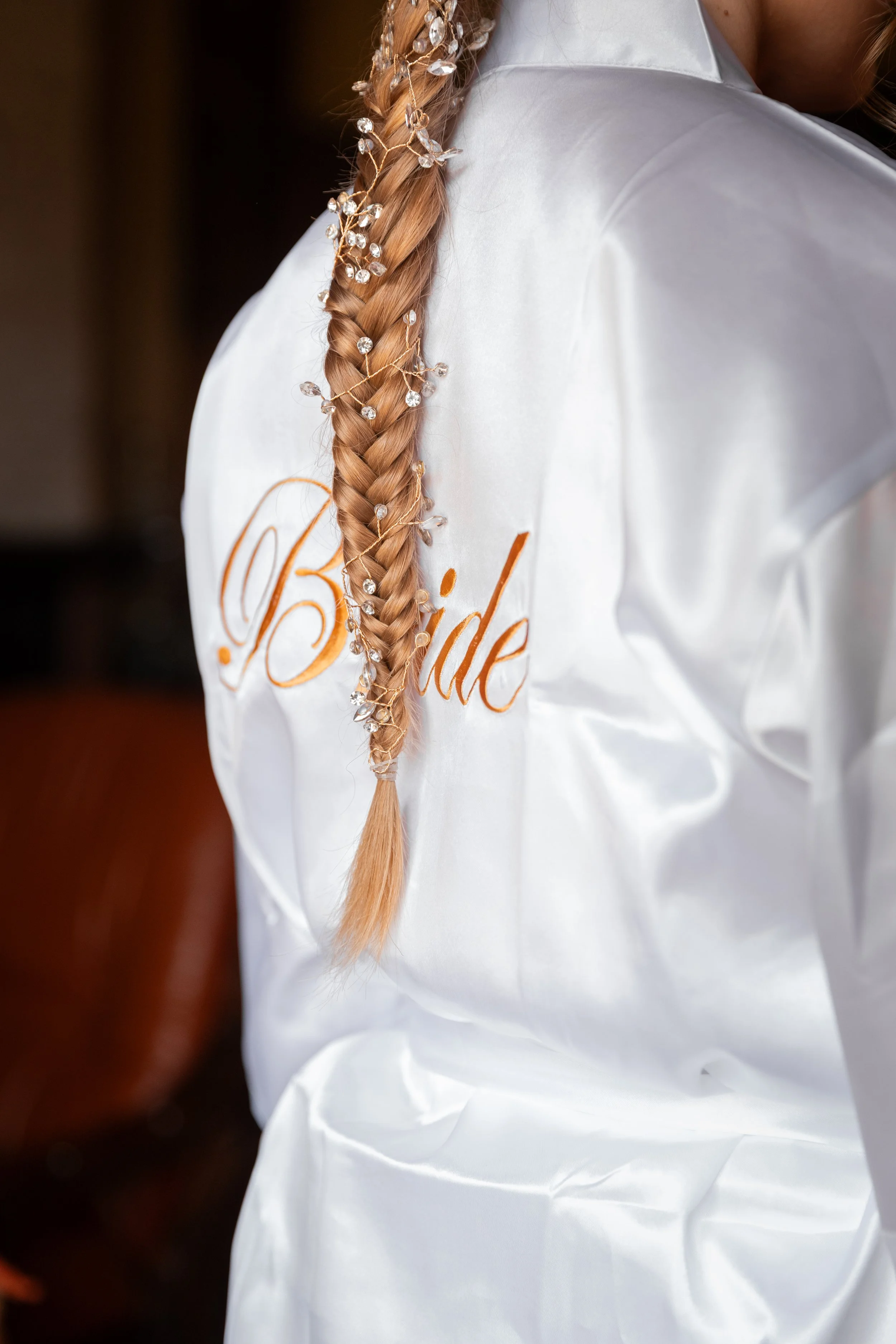 Coiffure d'une mariée avec une tresse décorée de perles et de rubans, portant une robe de satin blanc avec le mot 'Bride' en broderie orange.