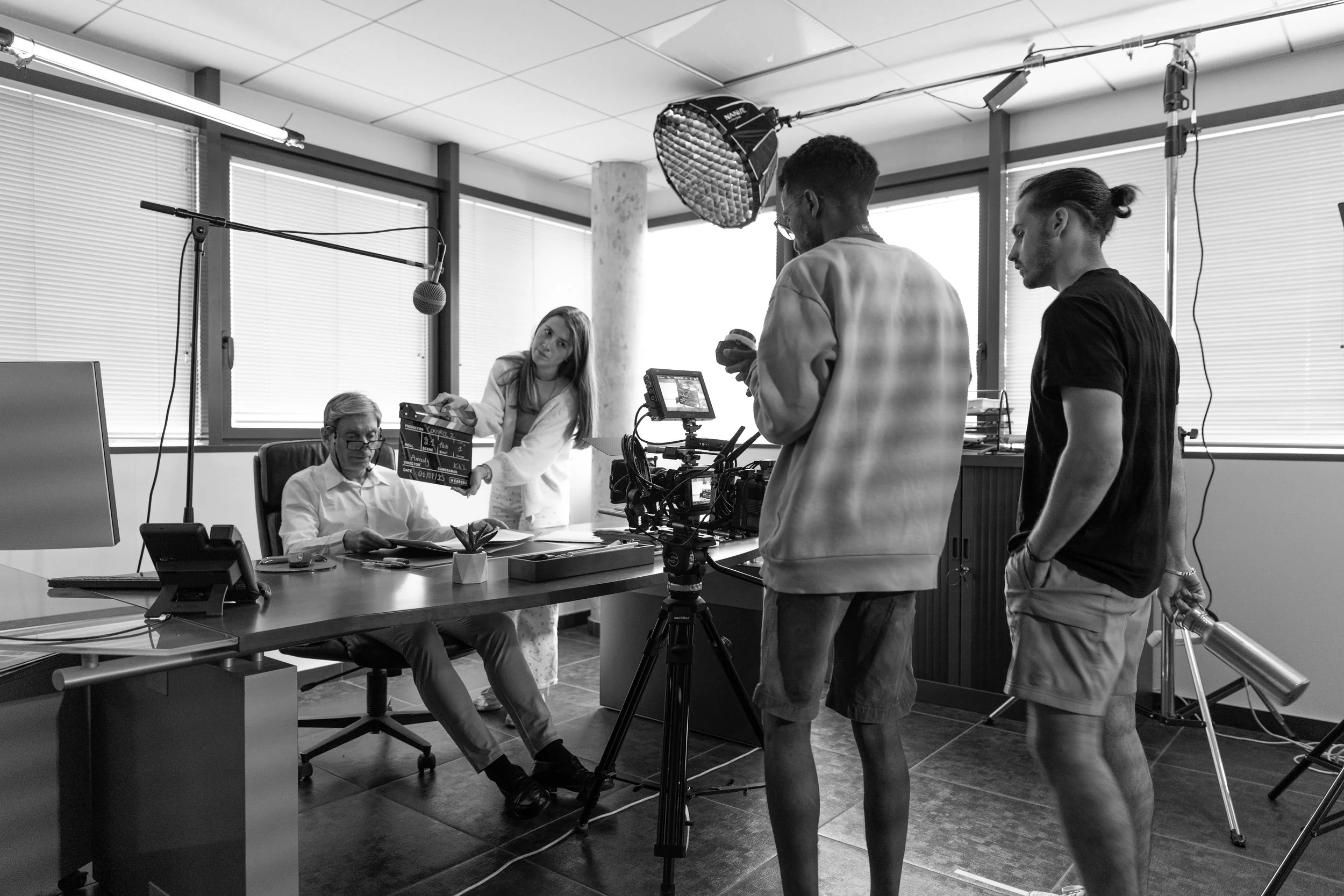 Une scène de tournage en intérieur, où une équipe de production filme un homme âgé assis à un bureau, avec une femme ajustant un clap de cinéma et deux hommes réglant du matériel technique.