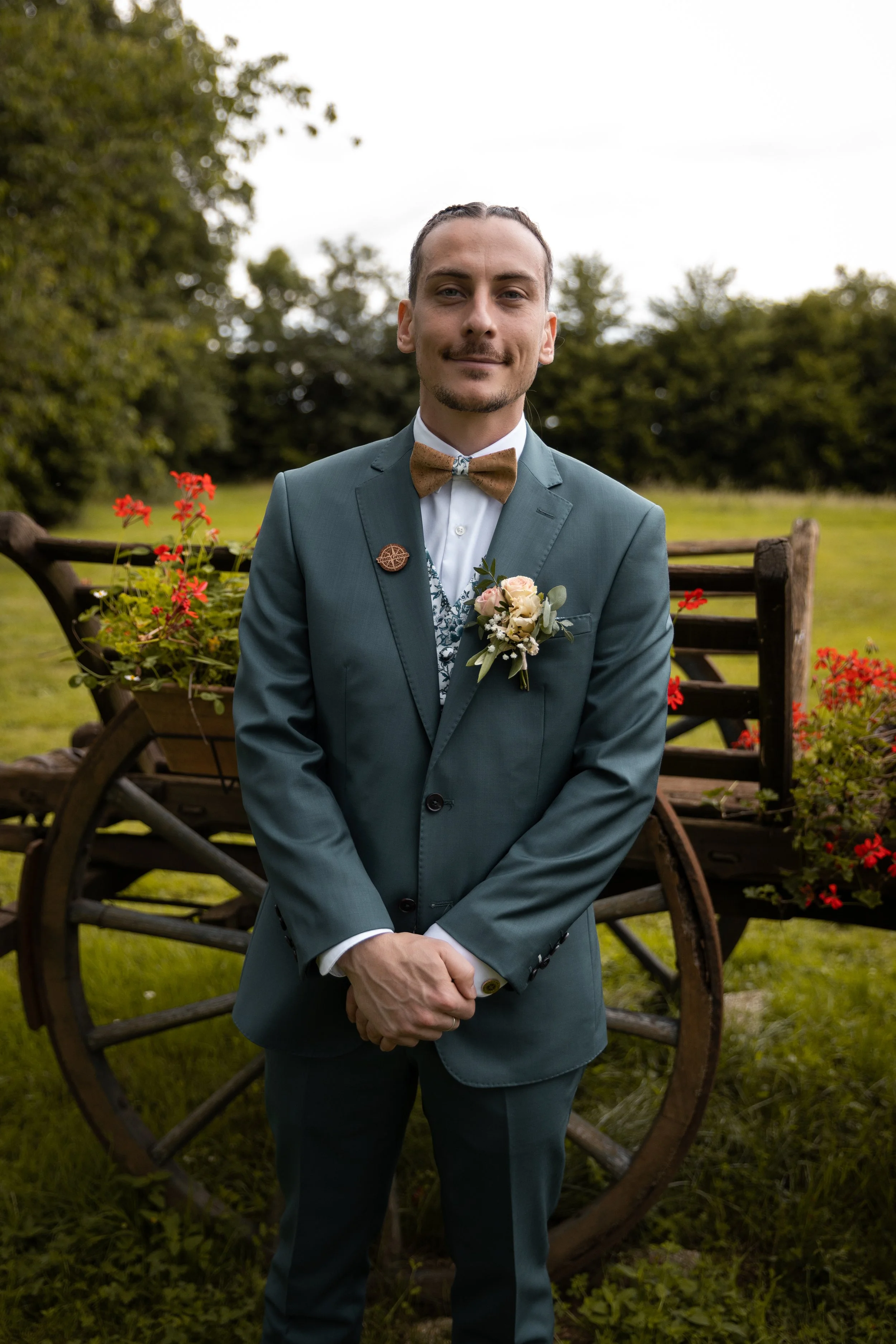 Un homme habillé en costume de mariage avec un nœud papillon en tweed, portant une boutonnière, se tenant devant un vieux chariot en bois décoré de fleurs rouges, dans un jardin vert.