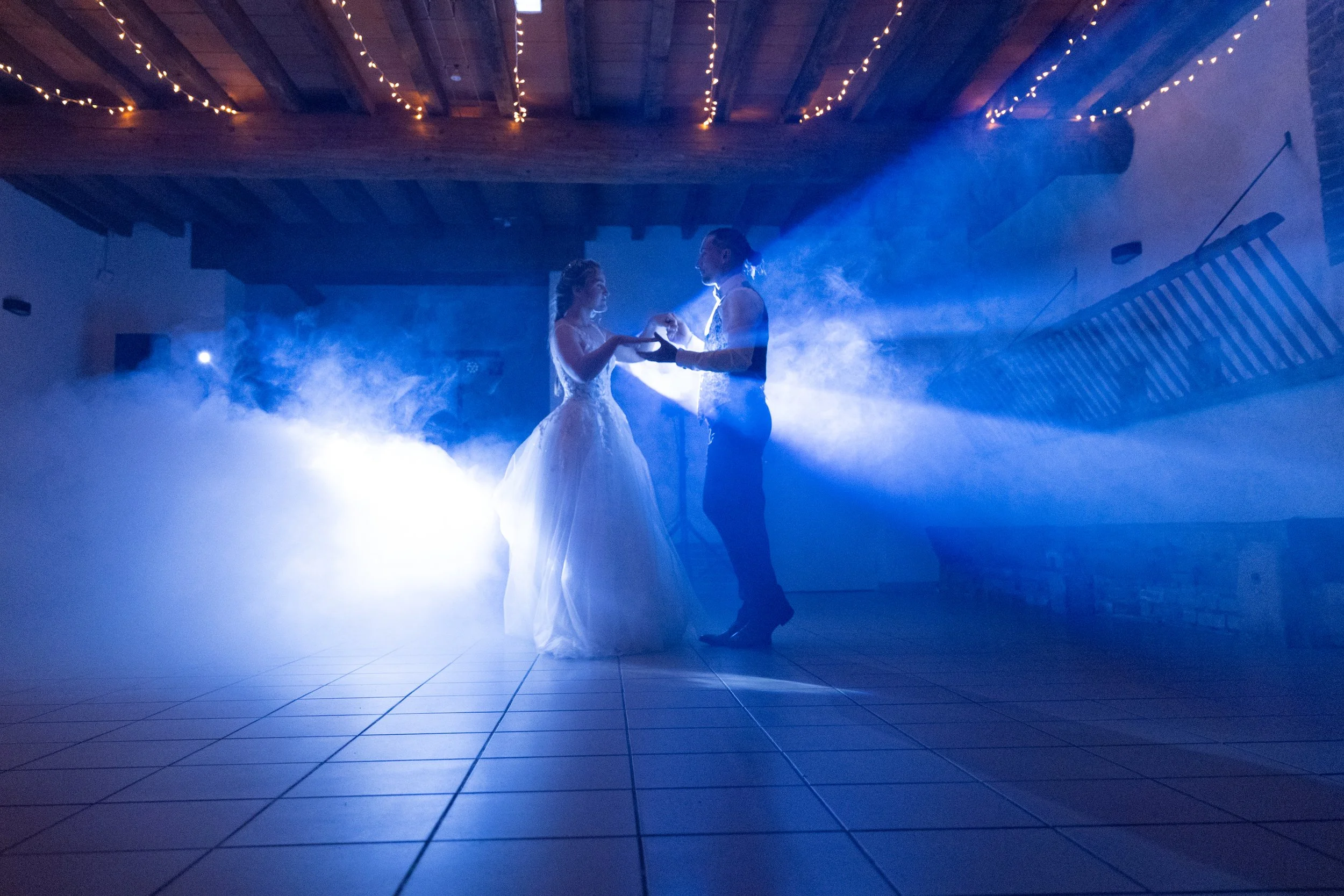Un couple danse lors de leur mariage, entouré de fumée et avec un éclairage bleu, dans une salle avec plafond en bois et guirlandes lumineuses.