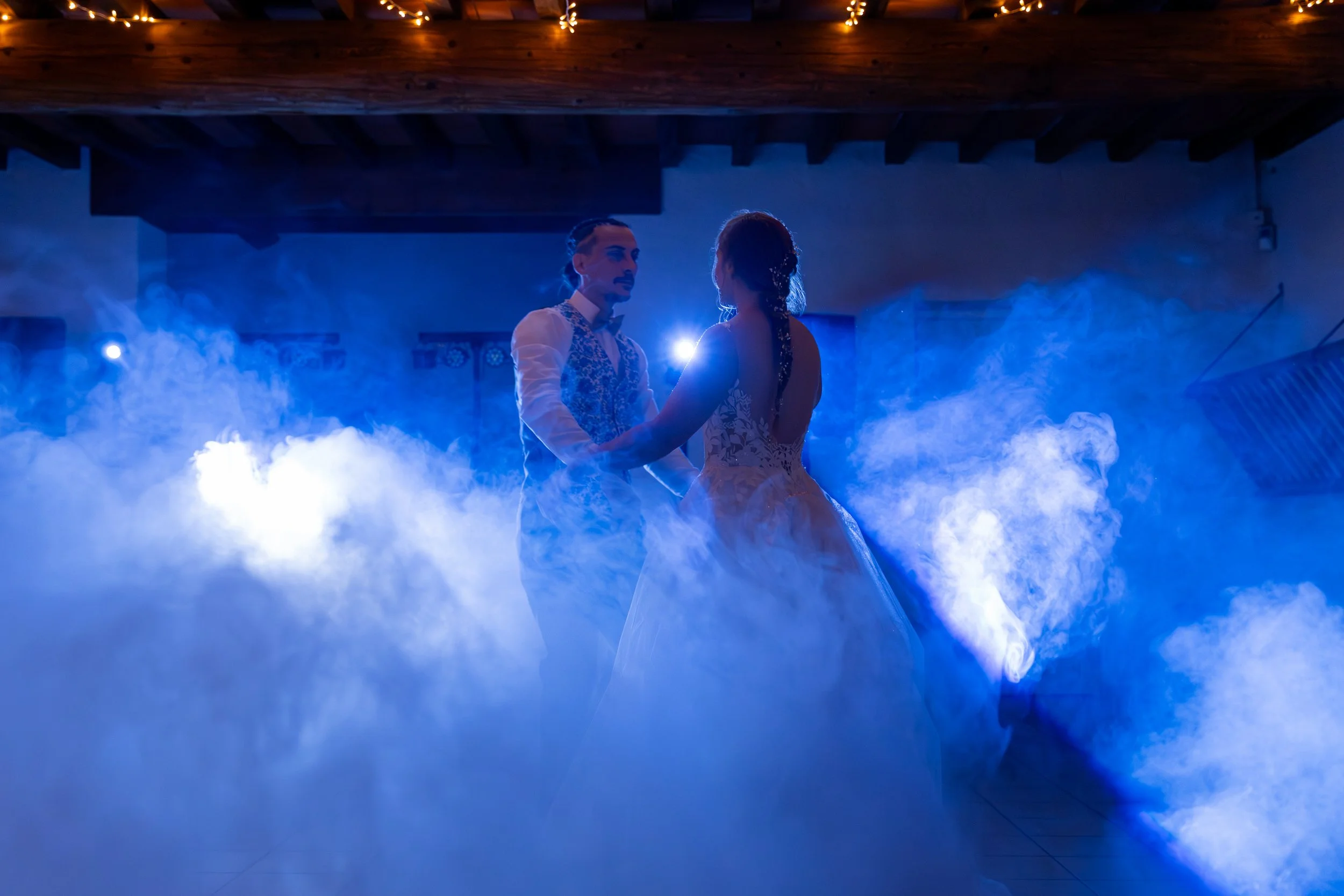 Un couple danse lors d'une cérémonie ou d'une réception, avec une ambiance mystérieuse créée par de la fumée et un éclairage bleu. La femme porte une robe de mariée et l'homme un costume veston.