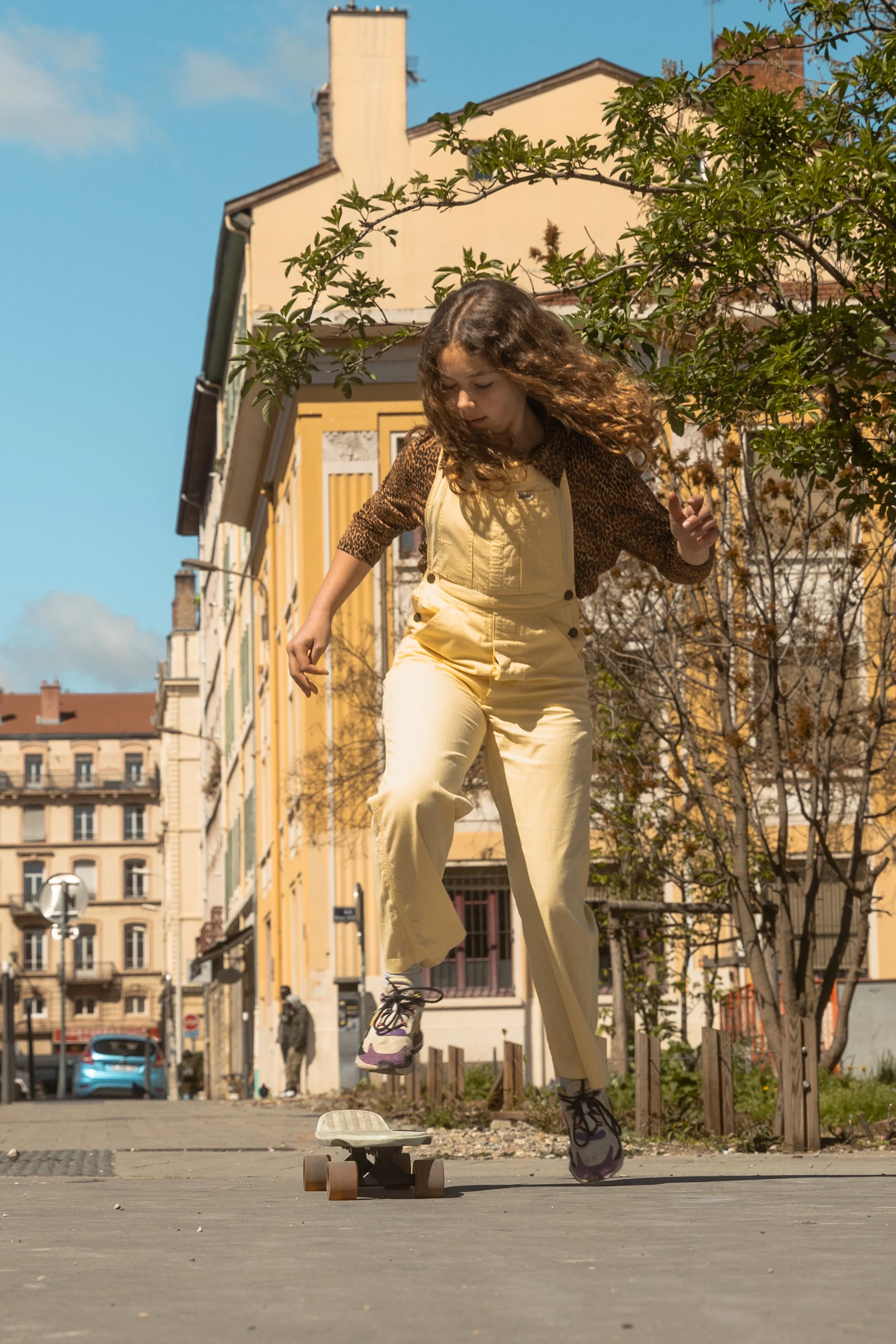 Une jeune fille qui fait du skateboard dans une rue urbaine ensoleillée, avec des bâtiments résidentiels en arrière-plan.